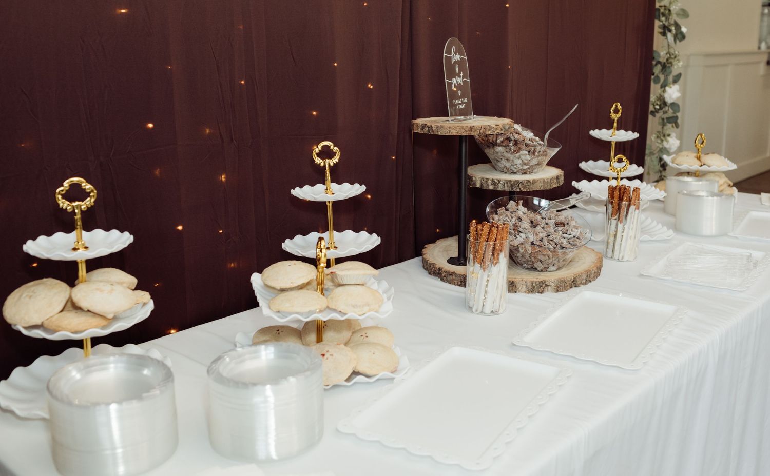Wedding dessert table with white tiered stands, cookies, chocolate-dipped pretzels, and rustic wood slice displays.