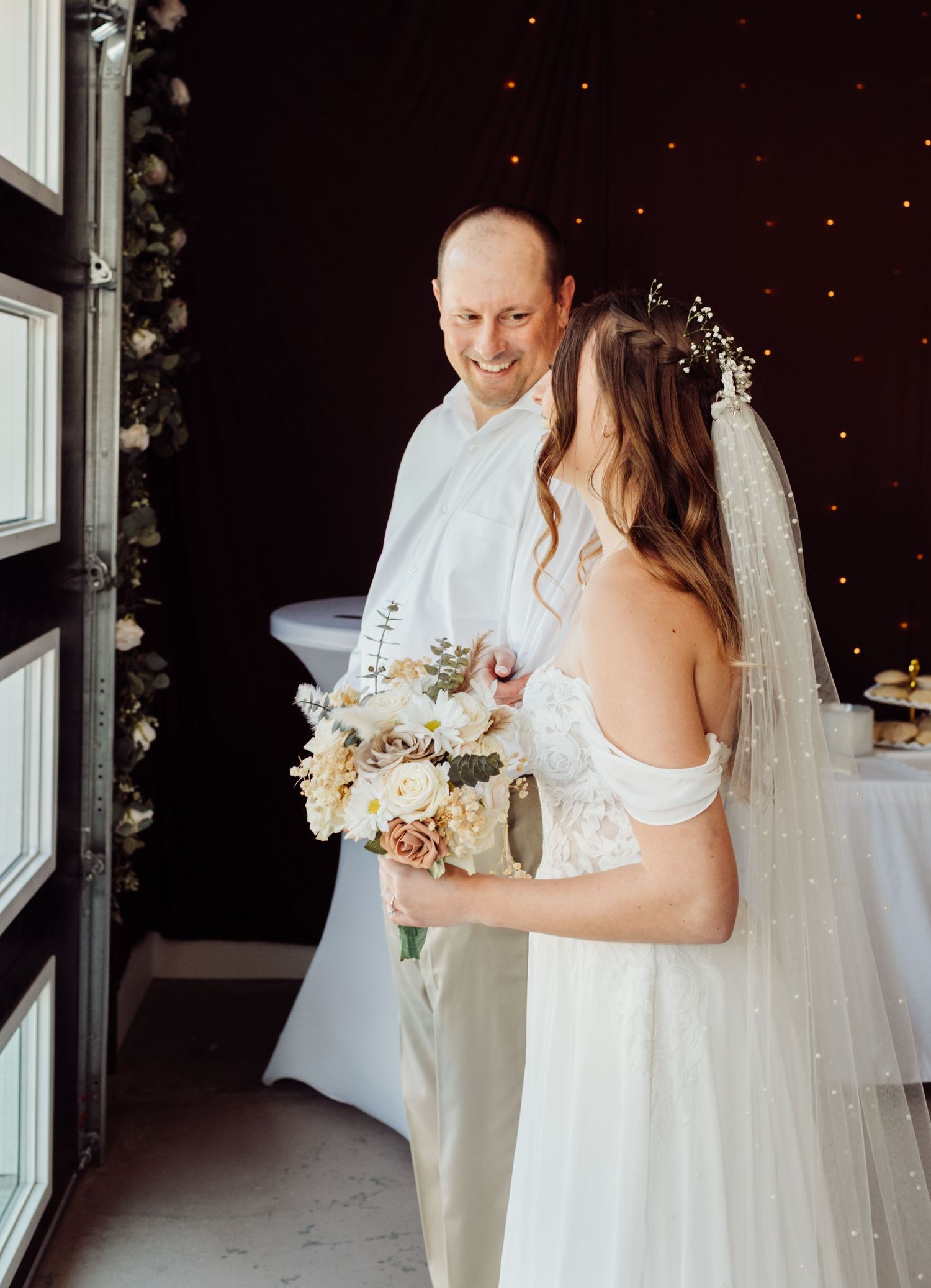 Bride in off-shoulder lace wedding gown holding a bouquet of white and blush flowers shares a moment with her father.