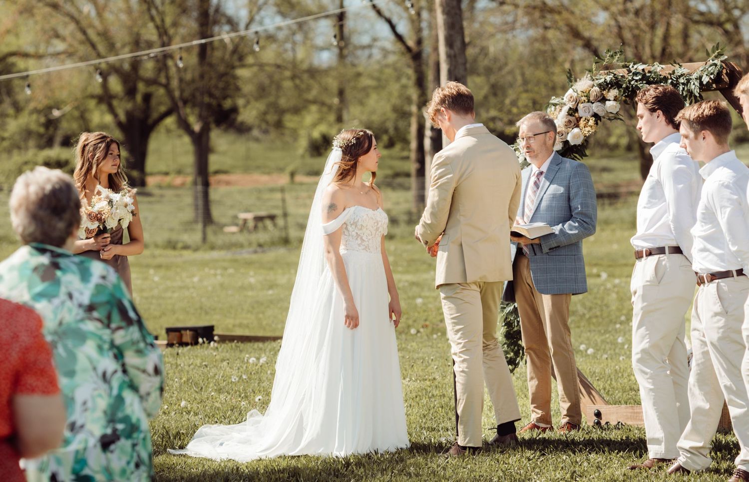 Outdoor wedding ceremony on green lawn with bride in white gown, groom in tan suit, and floral arch backdrop.