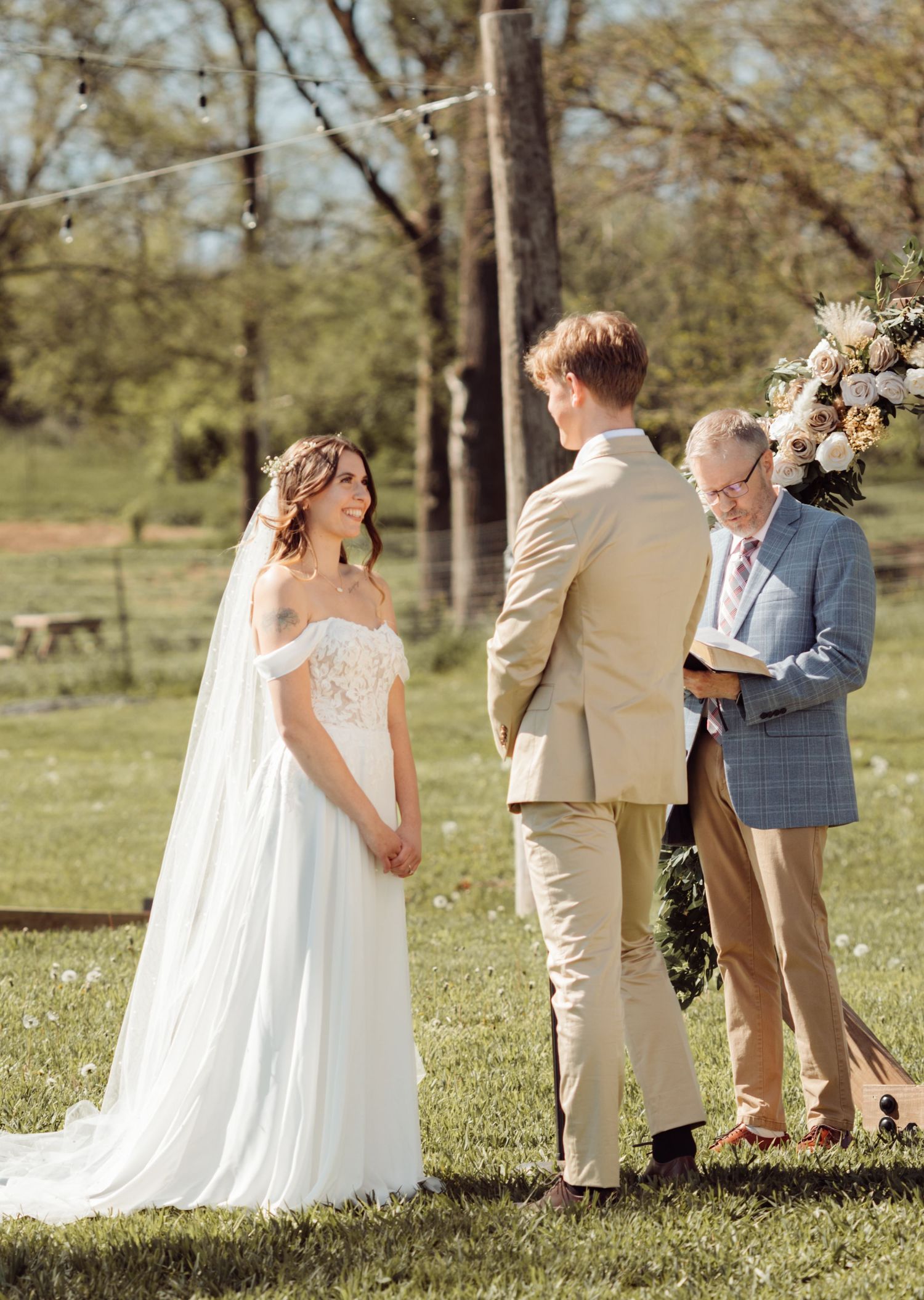 Bride in off-shoulder white gown with veil stands across from groom in tan suit during outdoor wedding ceremony.