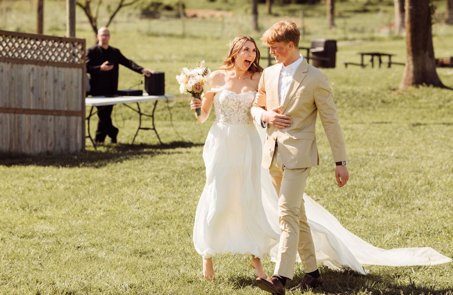 Joyful newlywed couple walking through green grass after outdoor wedding ceremony, bride holding white floral bouquet.