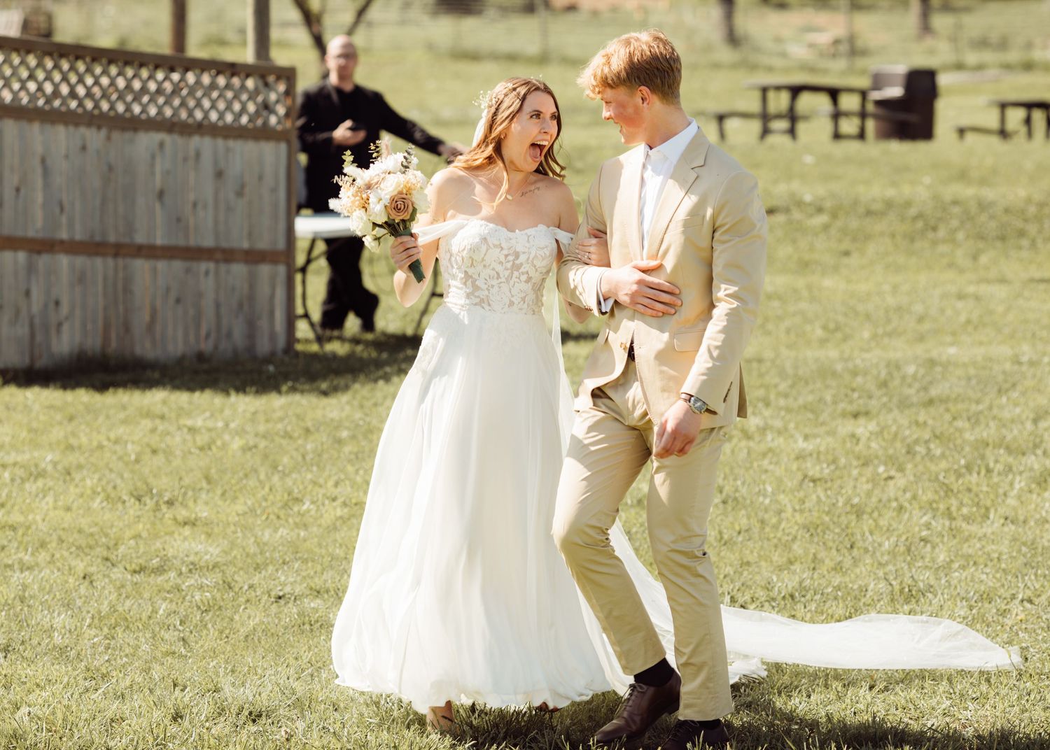Smiling bride in lace wedding gown and groom in tan suit strolling across green lawn after outdoor ceremony.
