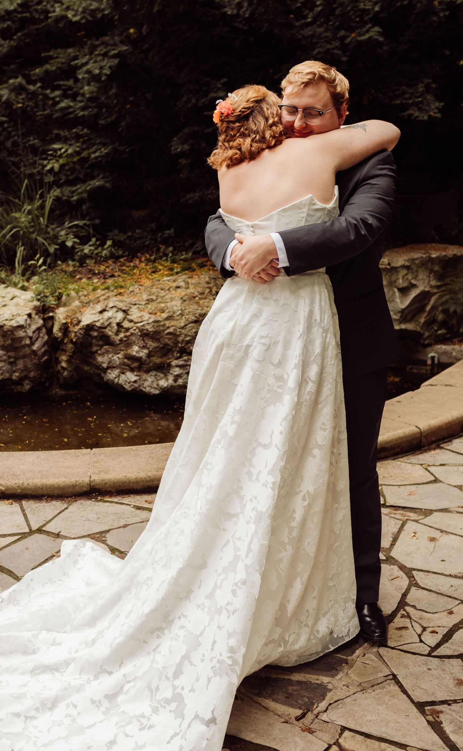 Bride and groom share a romantic first look hug on a stone patio near a waterfall.