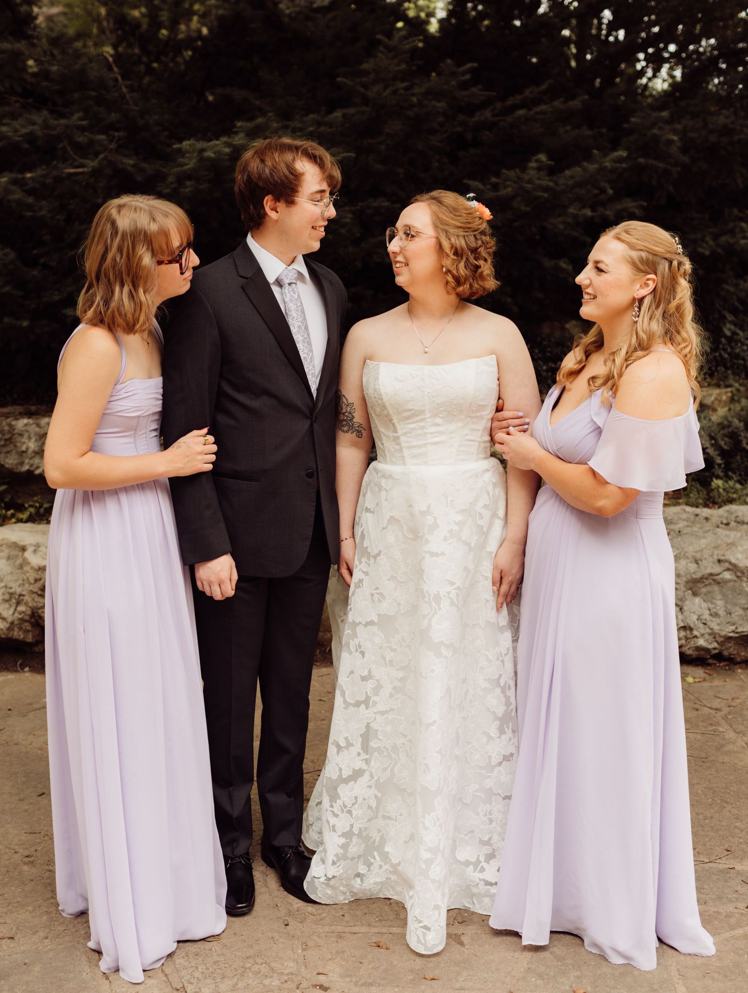 Bride in lace gown poses with groomsman and two bridesmaids in lavender dresses outdoors.