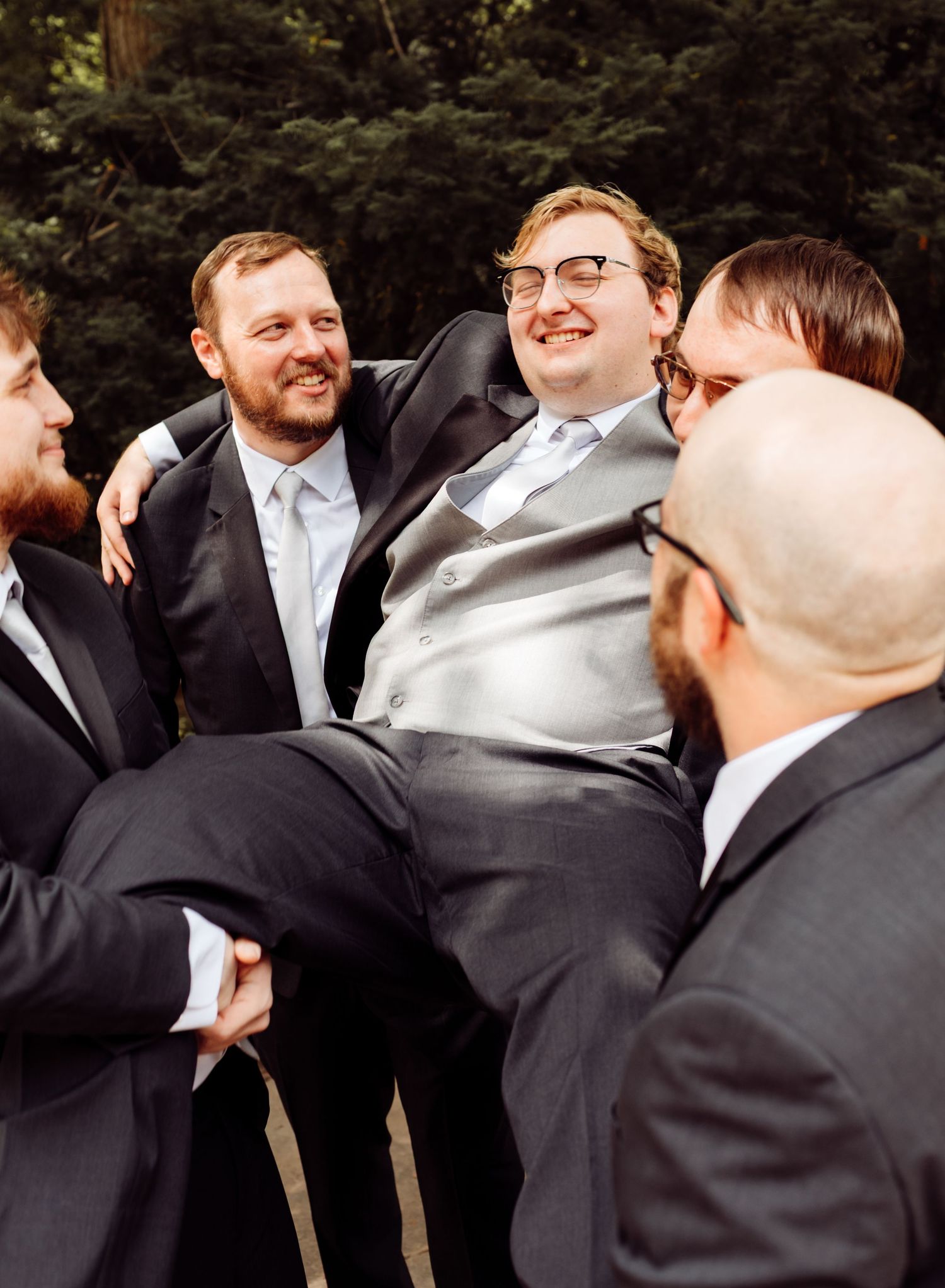 Groom is lifted by laughing groomsmen in dark suits during fun outdoor wedding photos.