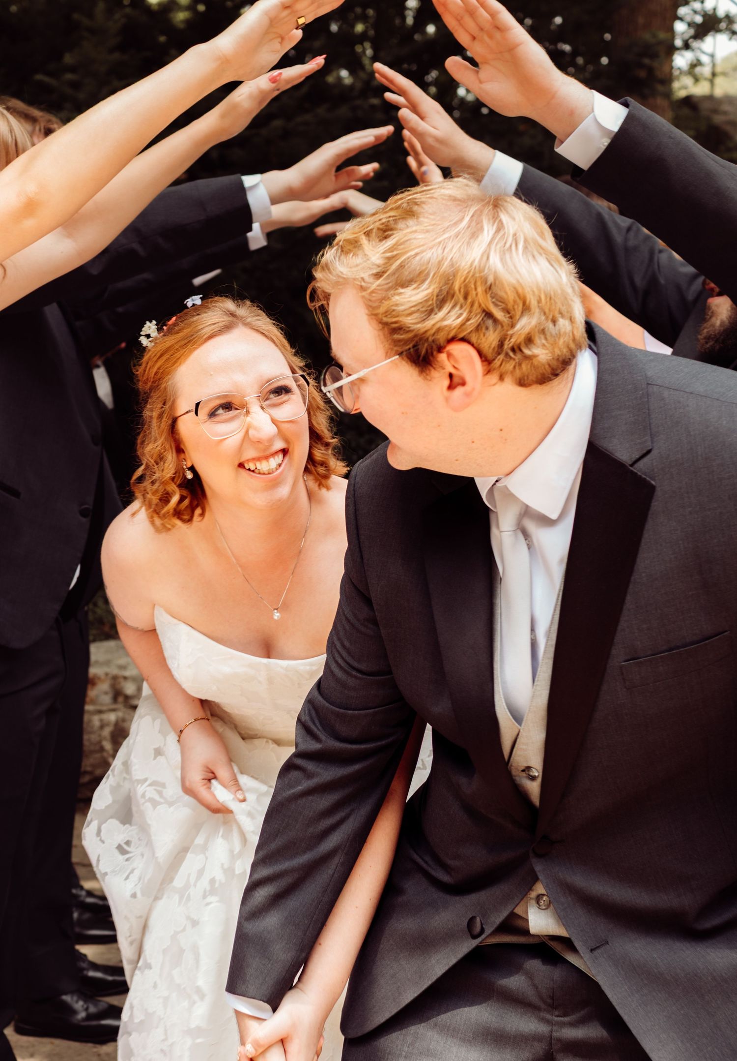 Bride and groom smile at each other while wedding party forms an arch overhead outdoors.