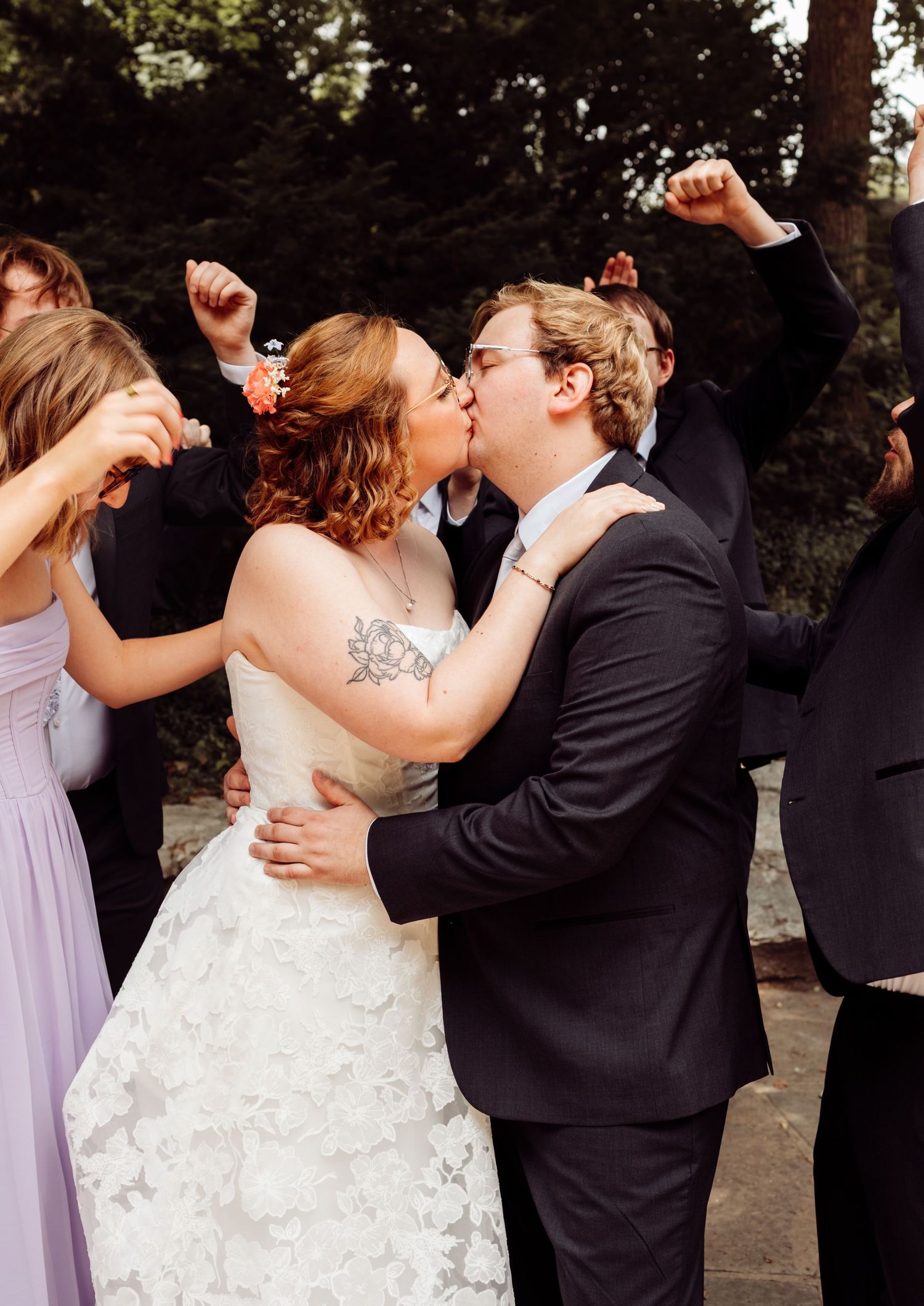 Newlyweds share a first kiss surrounded by cheering wedding party in lavender and black attire.