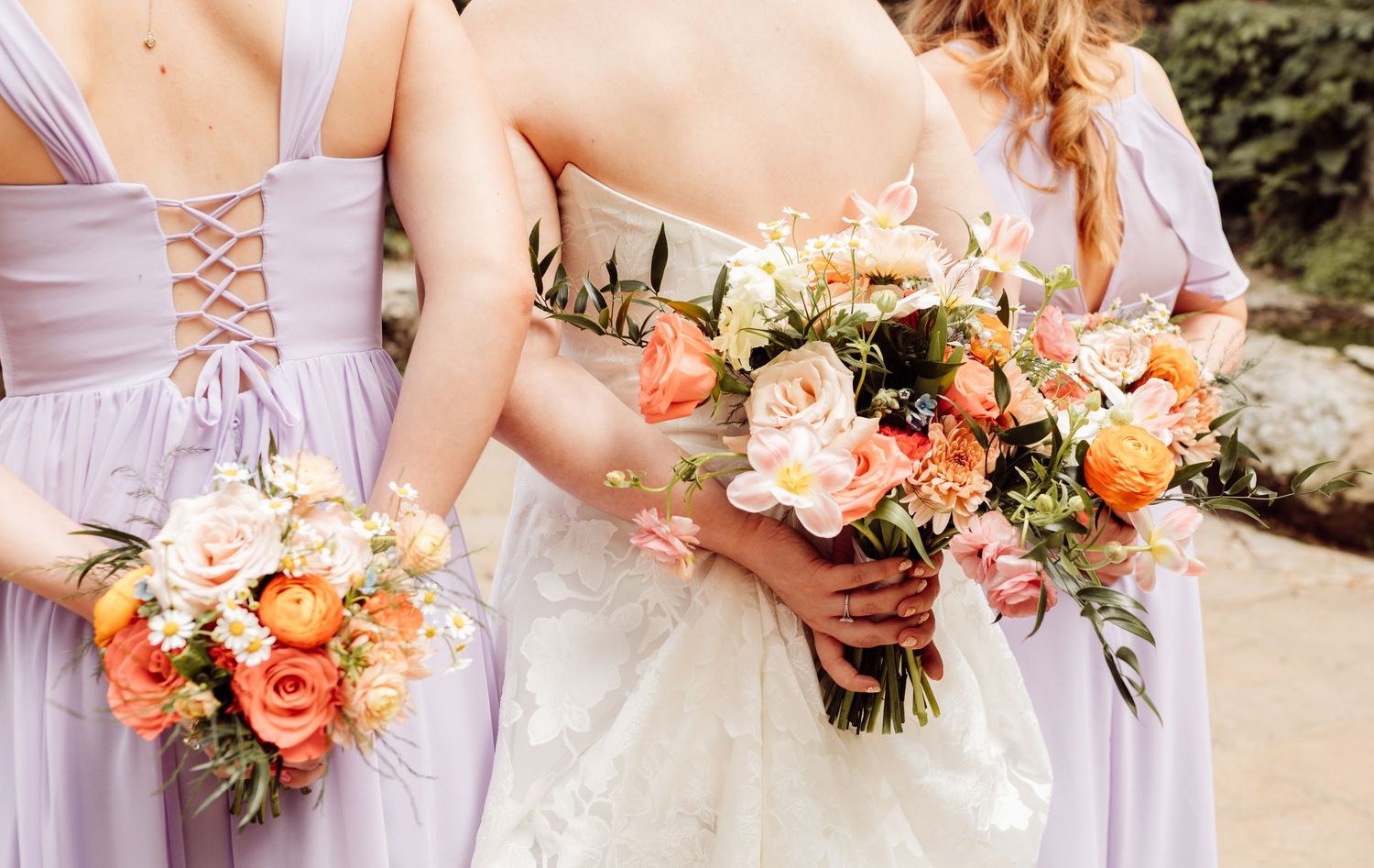 Bride and two bridesmaids in lavender gowns hold colorful peach and orange wedding bouquets.
