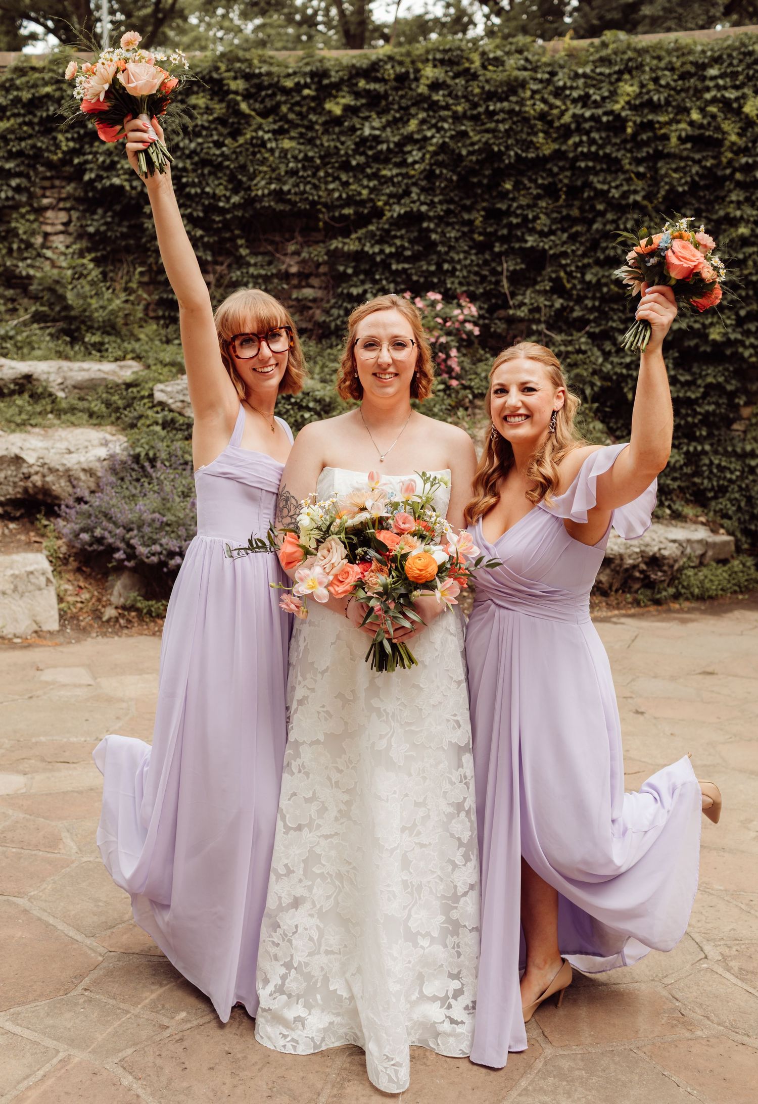 Bride in lace gown poses with two bridesmaids in lavender dresses raising bouquets outdoors.