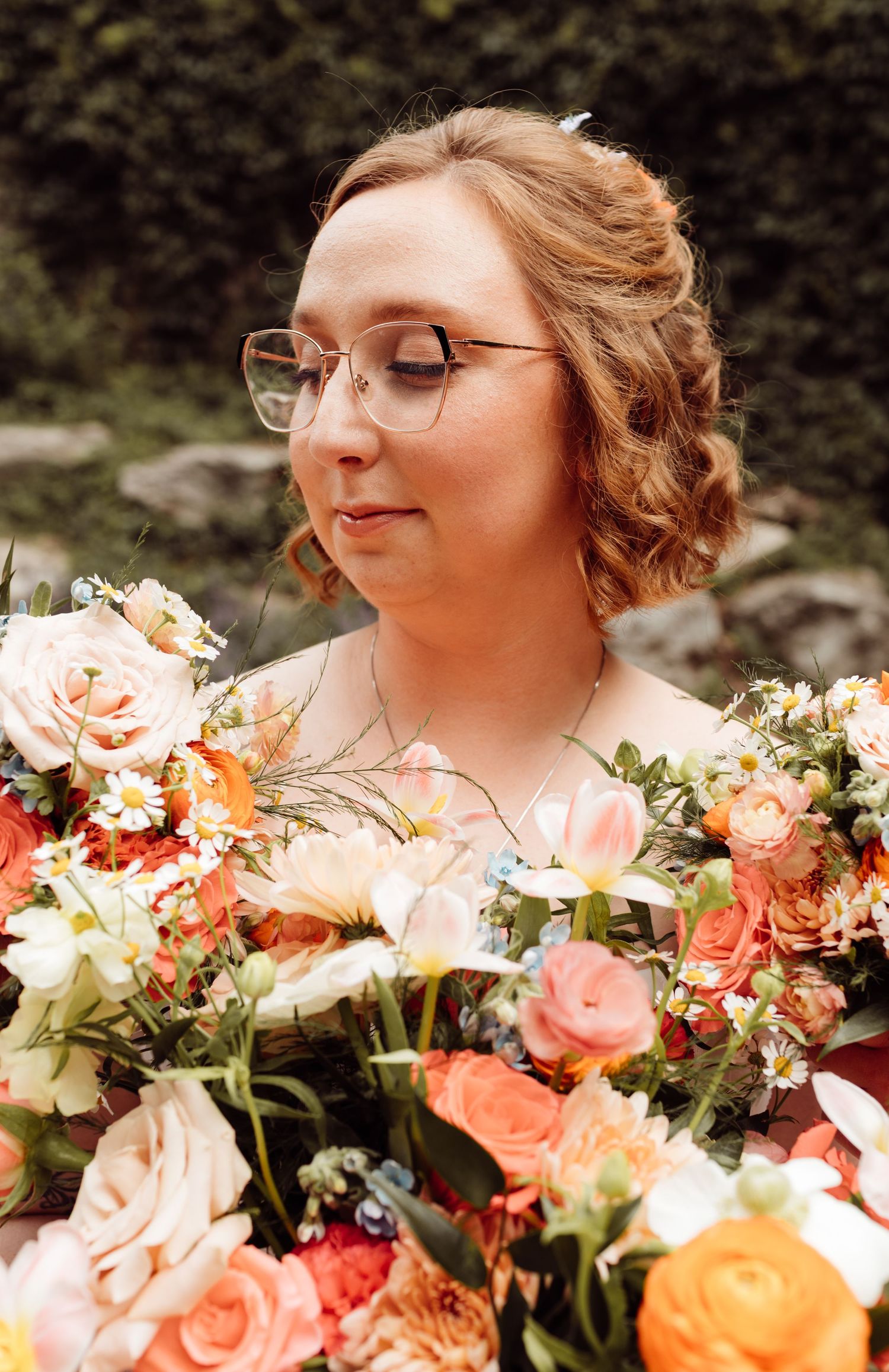 Bride with glasses and updo gazes down at vibrant orange and pink floral wedding bouquet.