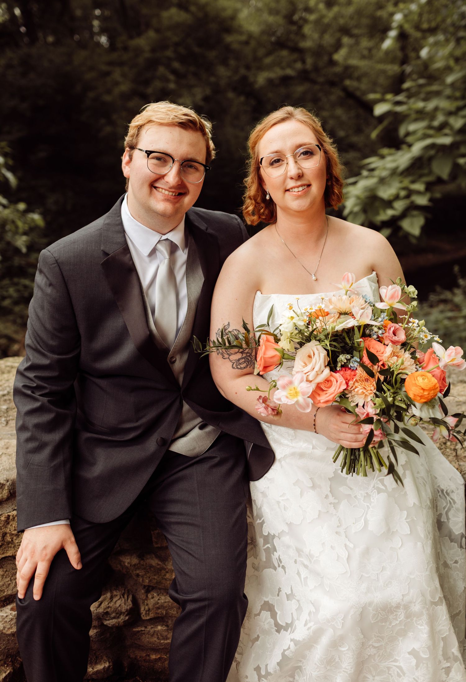 Smiling bride and groom sit together on stone wall surrounded by lush green foliage.