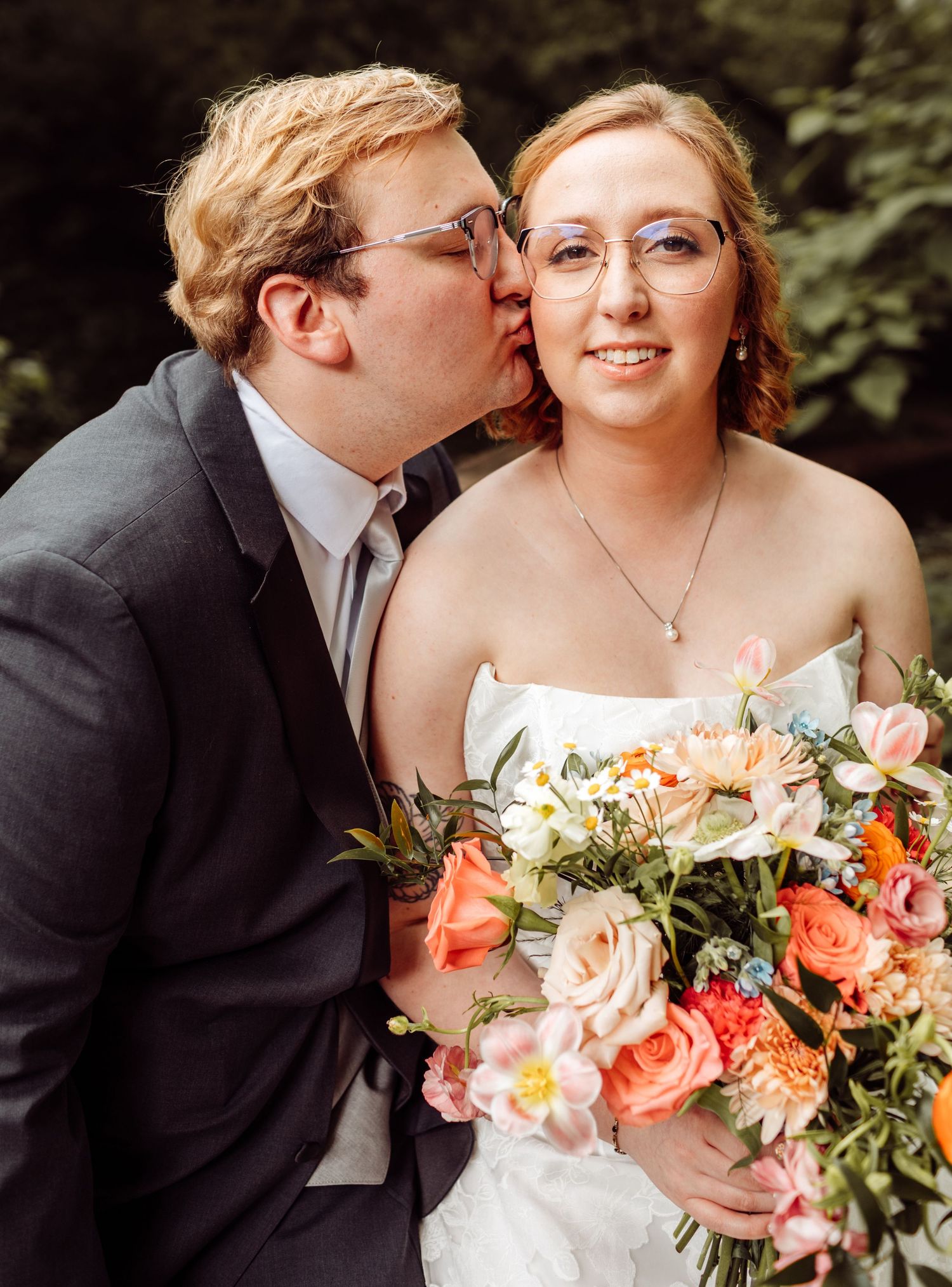 Groom kisses bride on cheek as she holds colorful peach and pink bouquet outdoors.