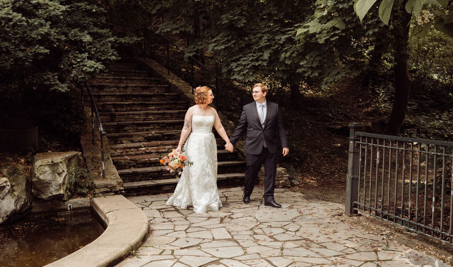 Bride and groom walk hand in hand on stone path near rustic staircase in garden.