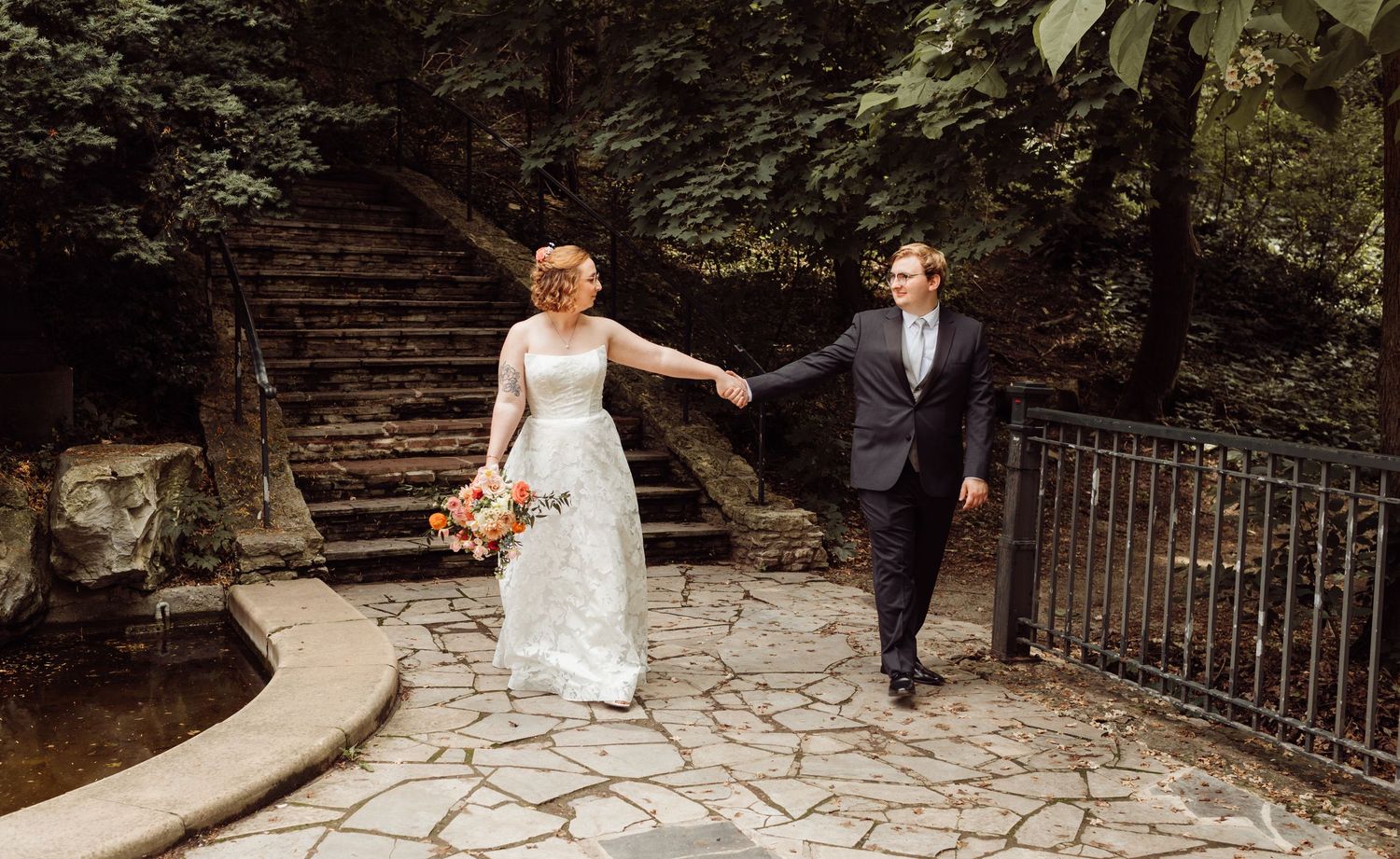 Bride leads groom by hand along stone garden path with lush green trees behind them.