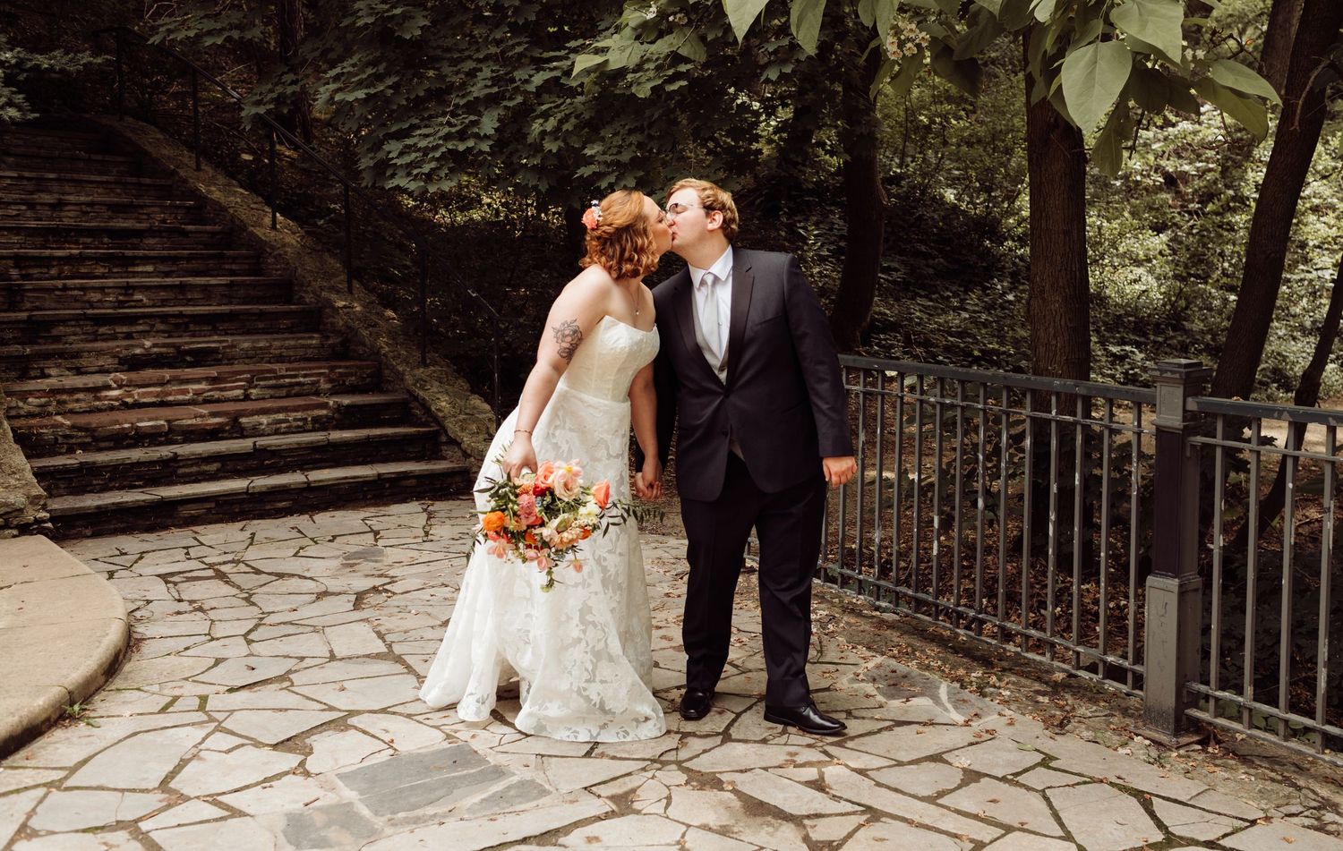 A bride and groom share a kiss on a stone patio surrounded by lush greenery and stairs during their wedding.