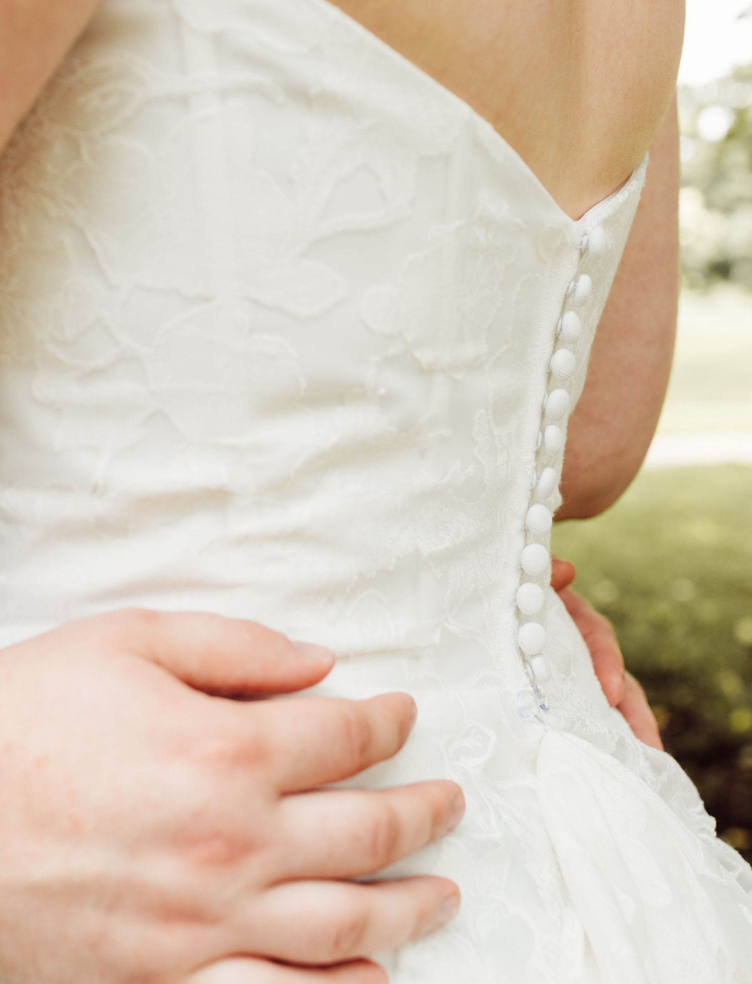 Close-up of a bride's white lace corset wedding dress with pearl buttons and a groom's hand on her waist.