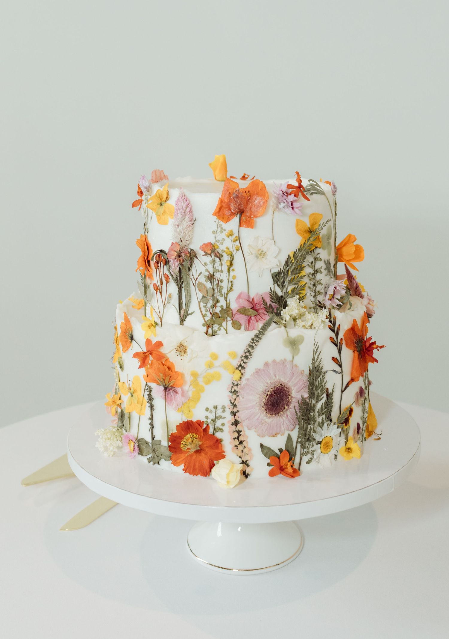 Elegant two-tier white wedding cake decorated with colorful pressed wildflowers on a white cake stand.