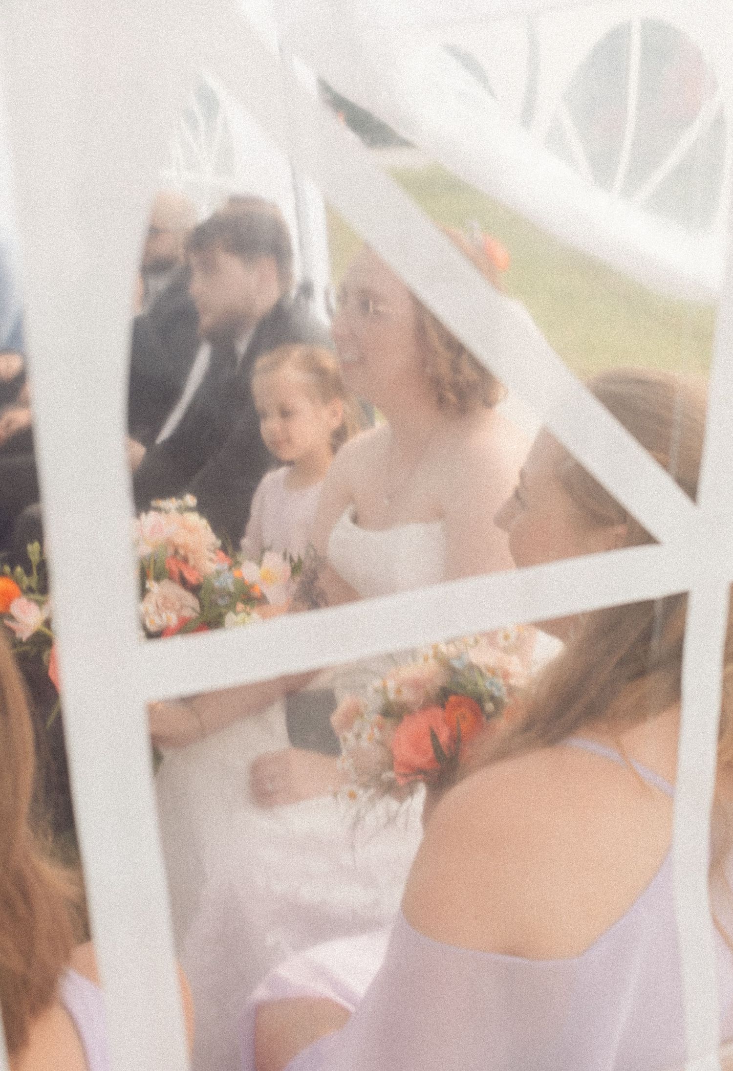 Bride holding a peach bouquet smiles at a flower girl during an outdoor wedding ceremony under a white tent.