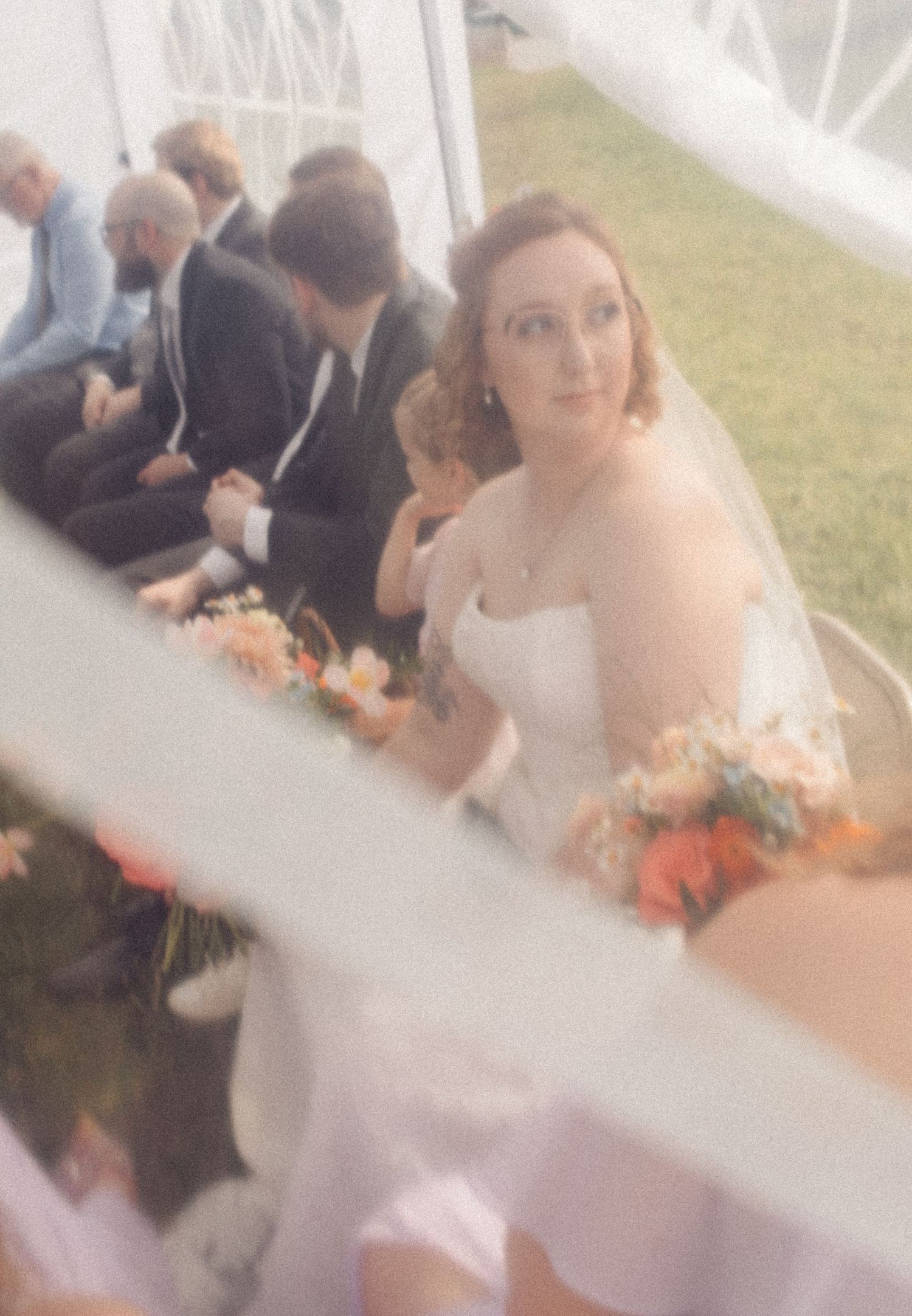 Bride in a white strapless gown with veil holds a peach bouquet while seated at an outdoor tent wedding ceremony.