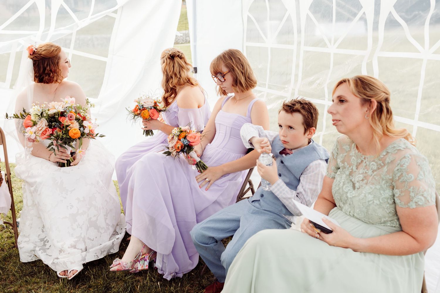 Bride and bridesmaids in lavender dresses sit together with bouquets during an outdoor wedding ceremony under a tent.