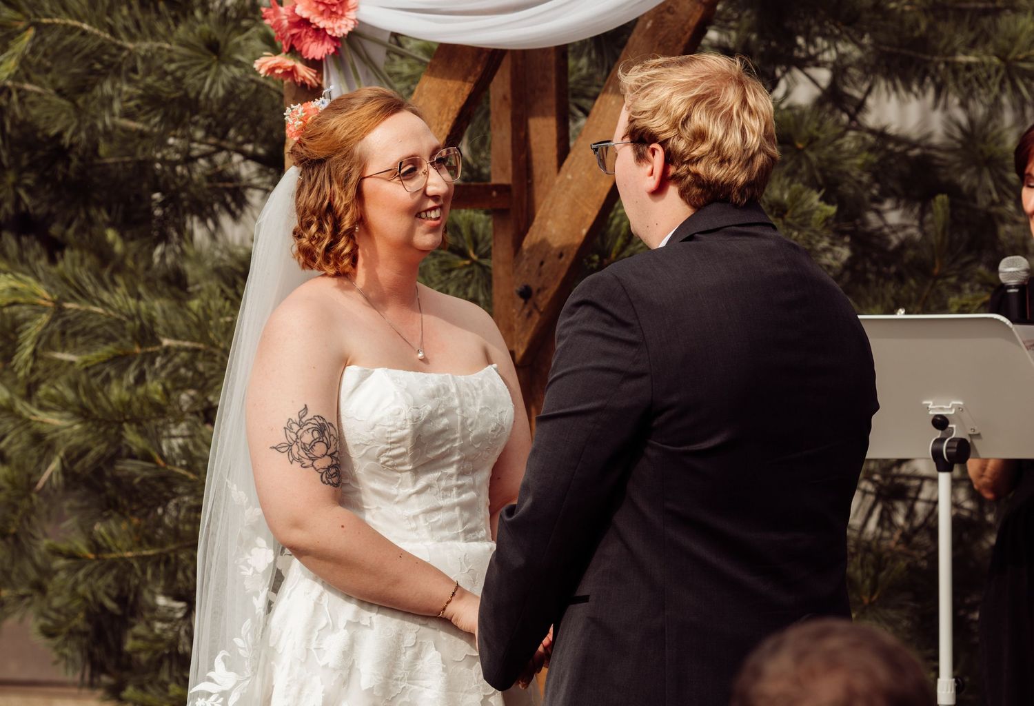Bride with a rose tattoo and veil smiles at her groom during outdoor wedding vows beneath a wooden arch.