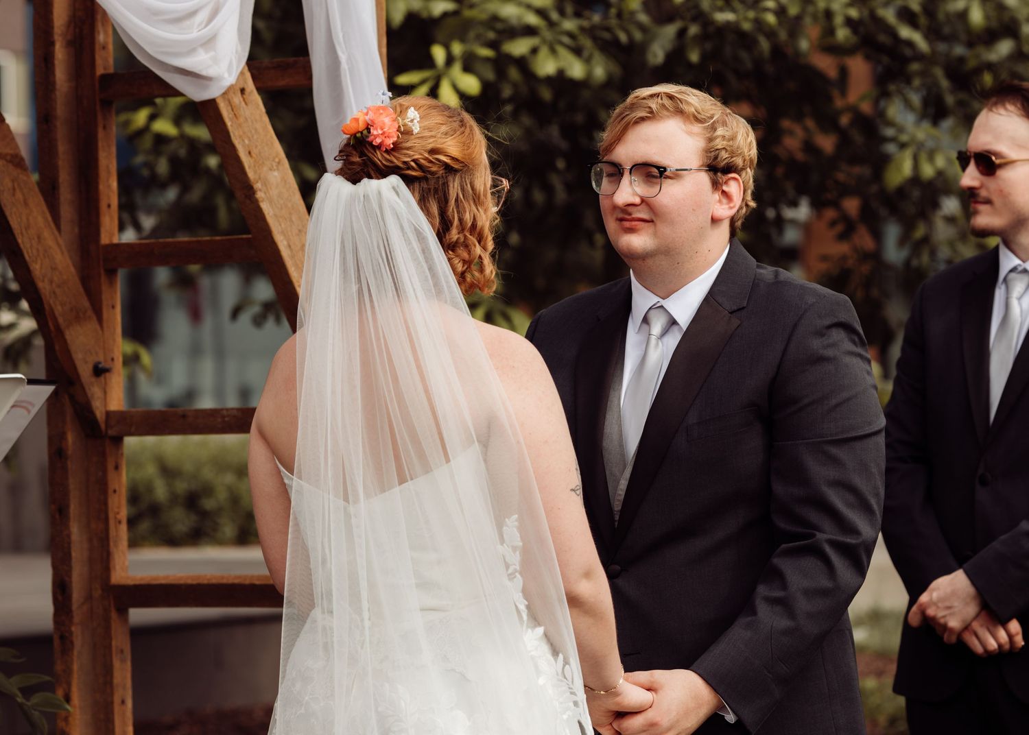 Groom in a black suit smiles warmly at his bride during outdoor wedding ceremony beneath a draped wooden arch.