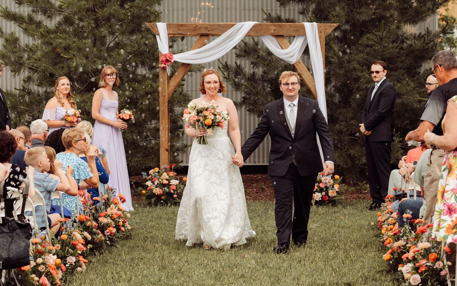 Bride and groom walk joyfully down the aisle after their outdoor wedding ceremony lined with colorful floral arrangements.