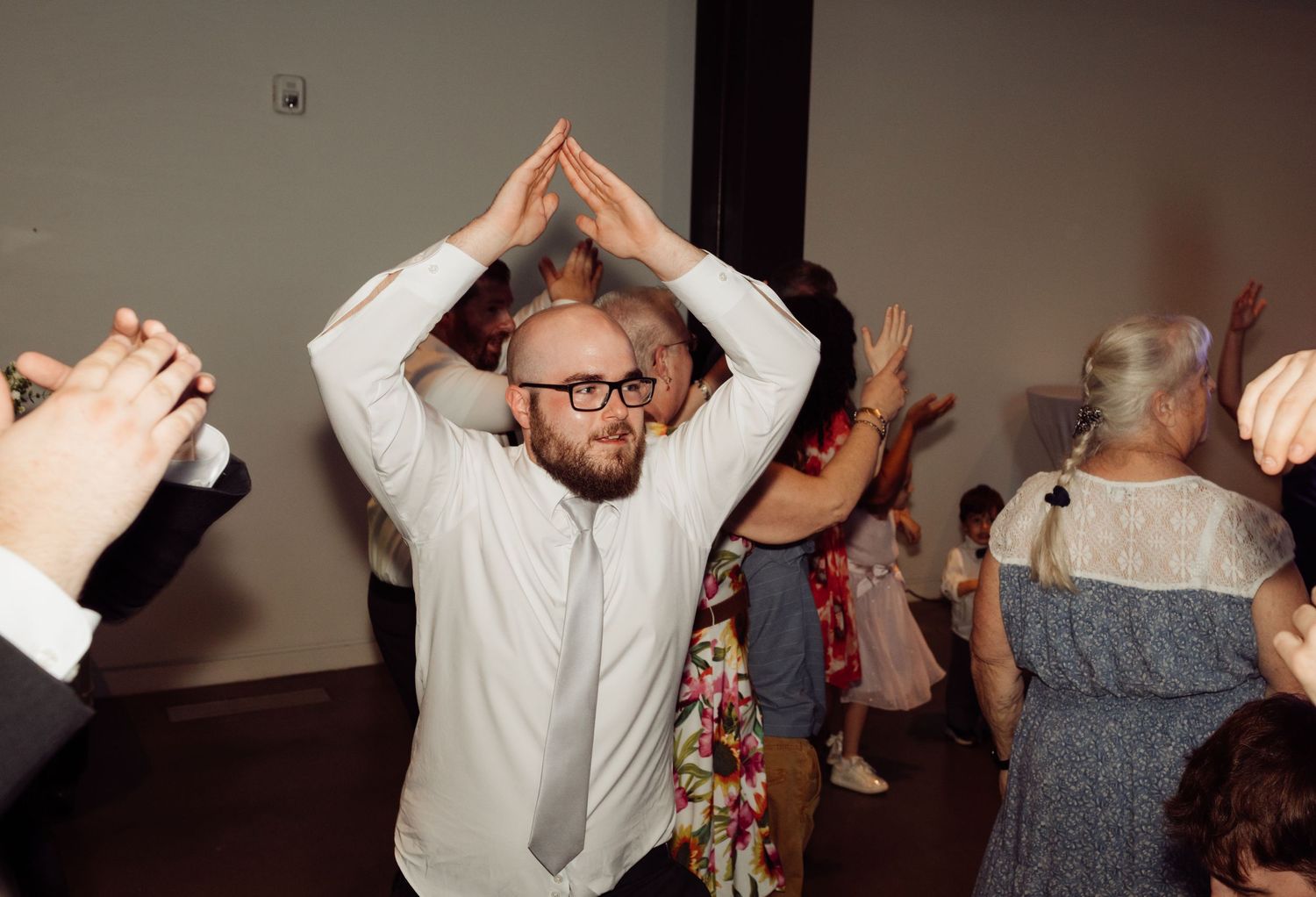 Bald man with glasses dancing enthusiastically with arms raised at a wedding.