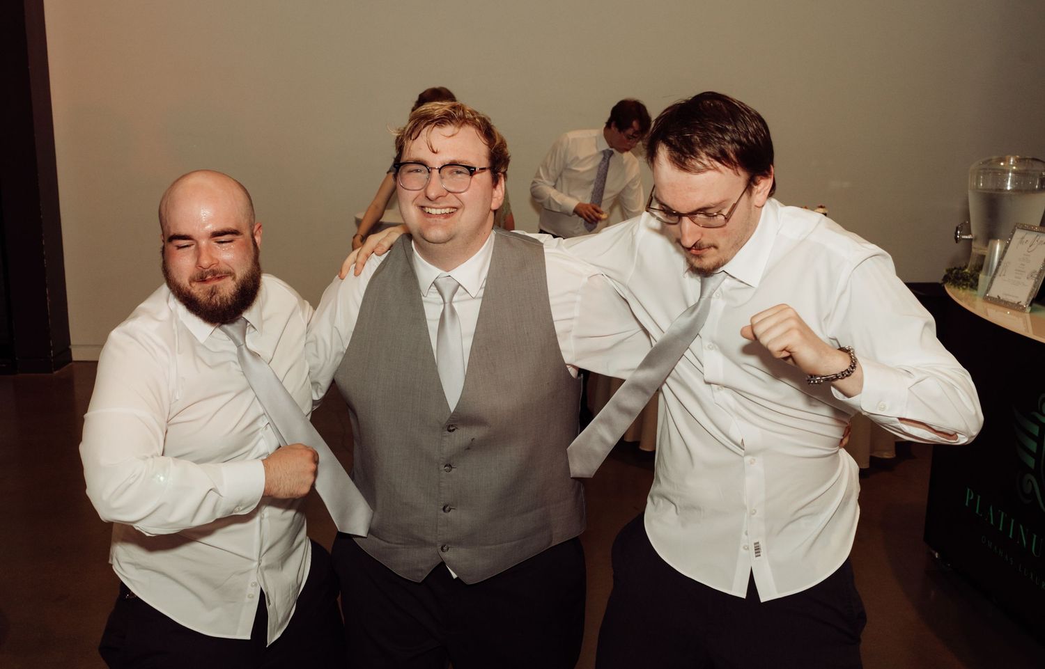 Three men in dress shirts pose playfully at a wedding reception, making fist gestures and smiling.