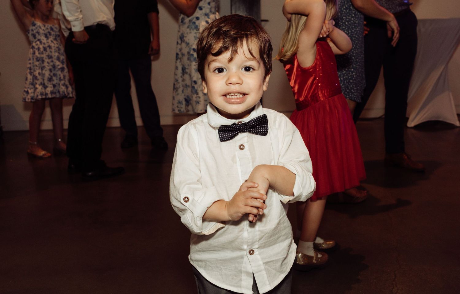 Adorable toddler boy in white shirt and bow tie smiles on a wedding dance floor.