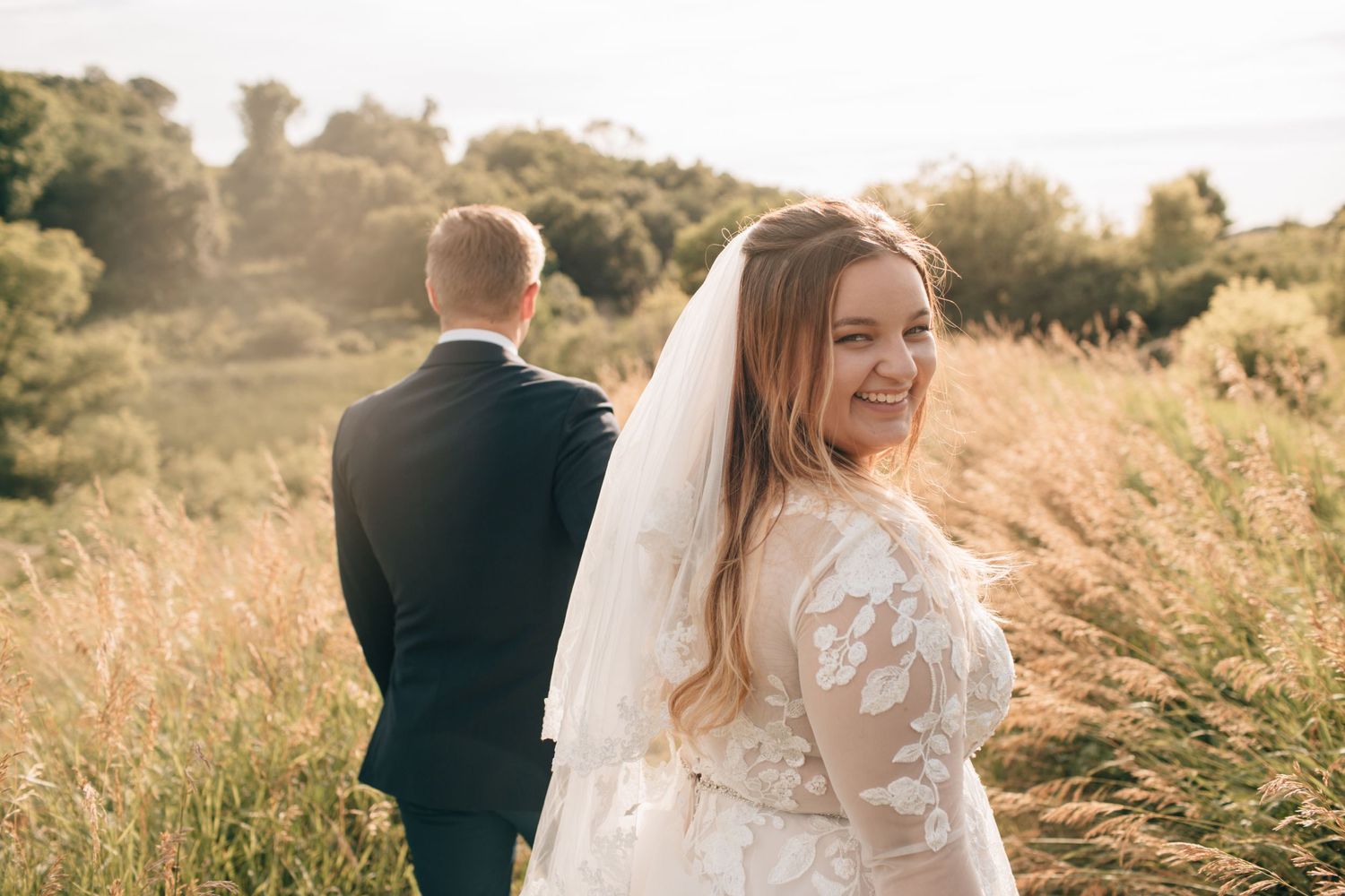 Bride in floral lace gown and veil smiles back at camera while groom stands in golden field.