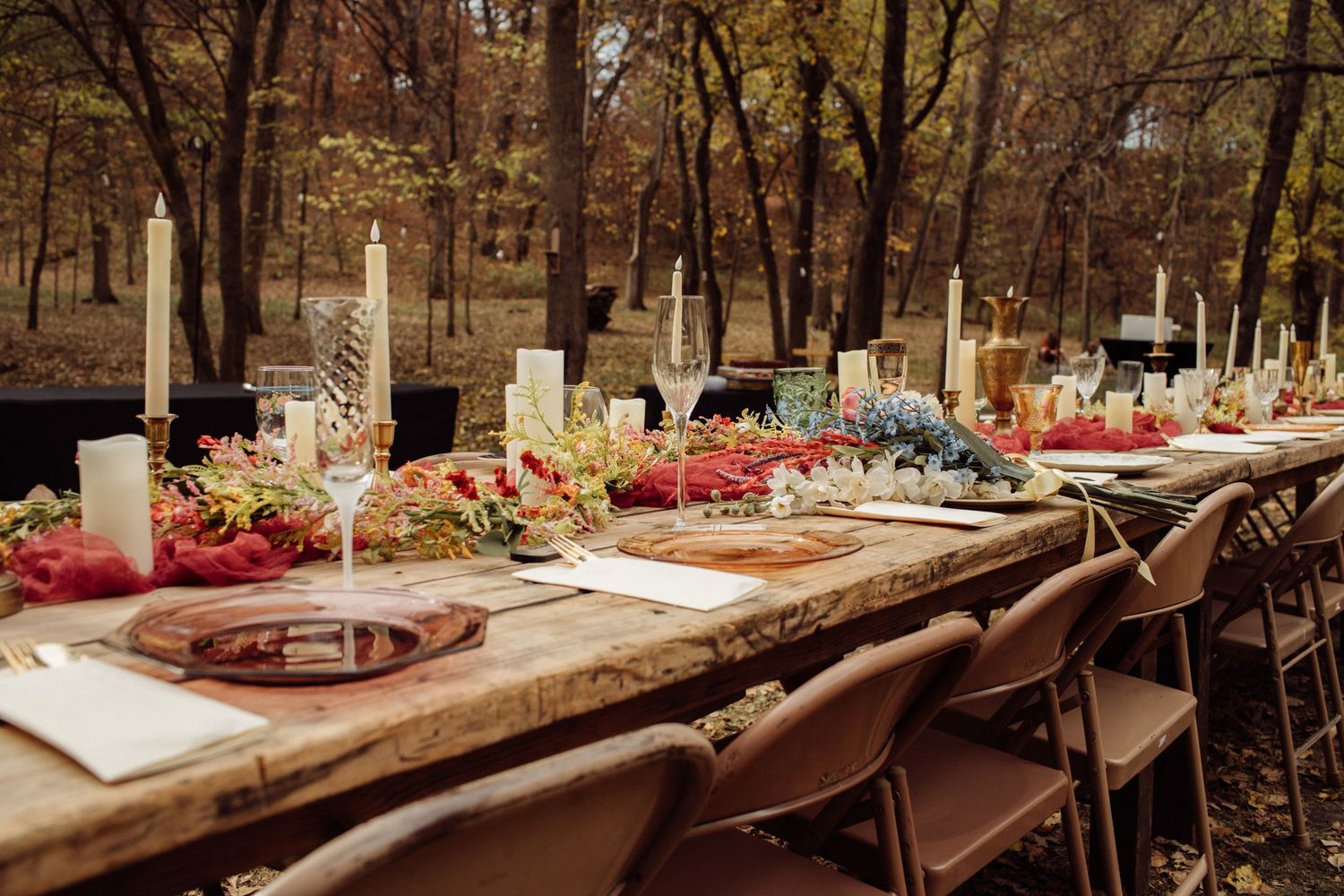 Elegant outdoor woodland wedding reception table set with candles, colorful florals, and rustic wood decor.