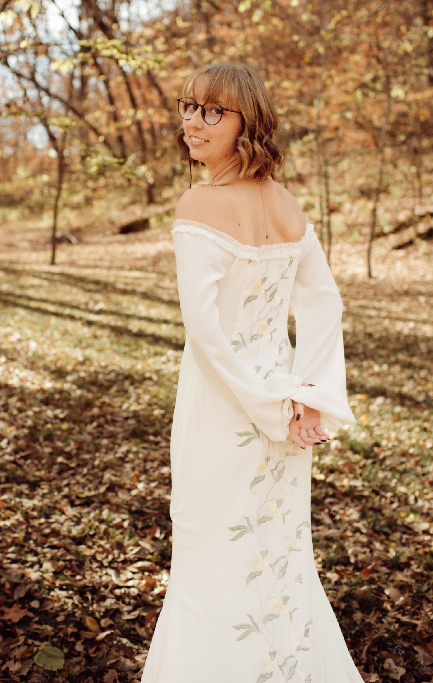 Bride in off-shoulder white gown with floral embroidery posing in sunlit autumn forest.