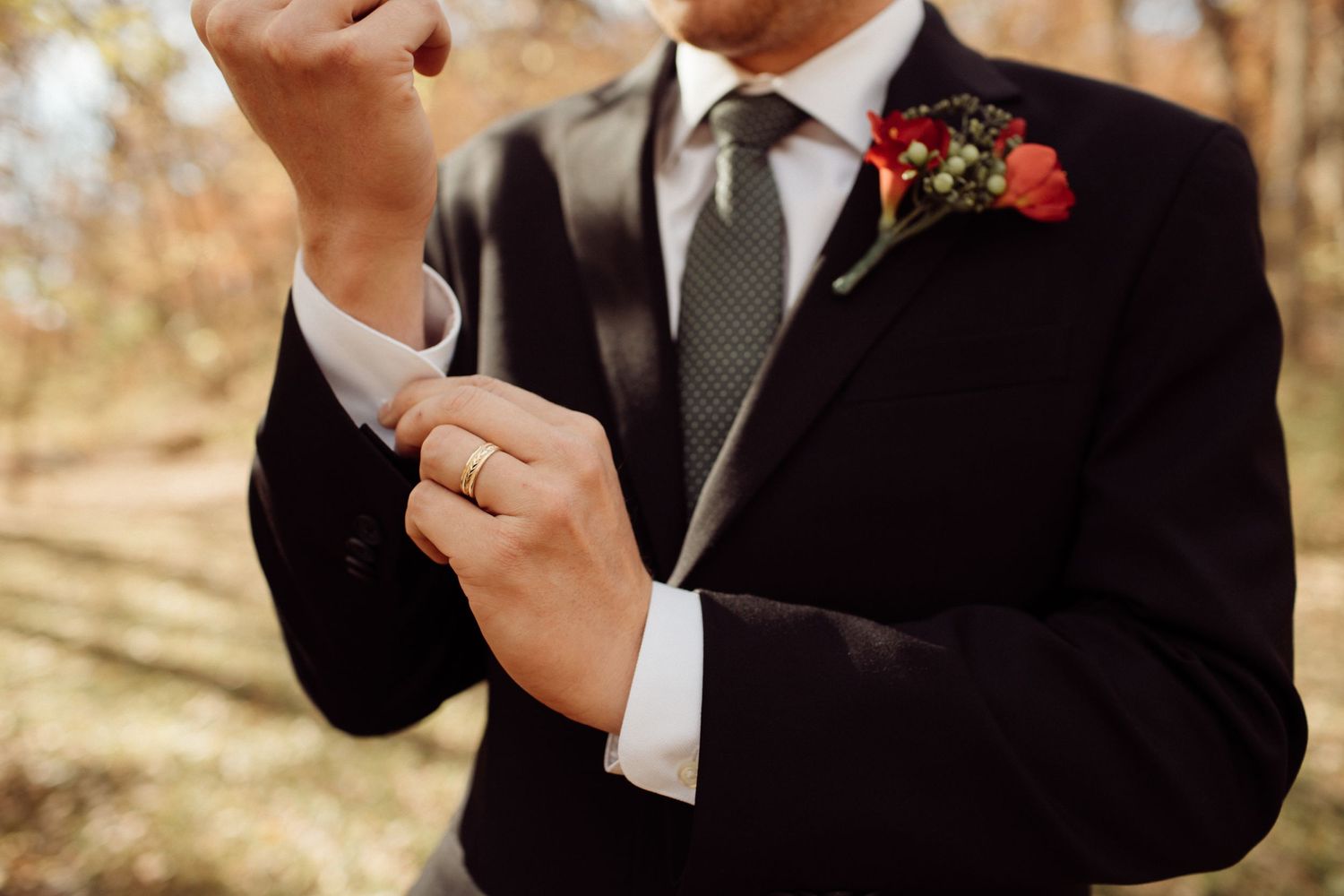 Groom in black suit adjusting cufflinks with red floral boutonniere and gold wedding band.