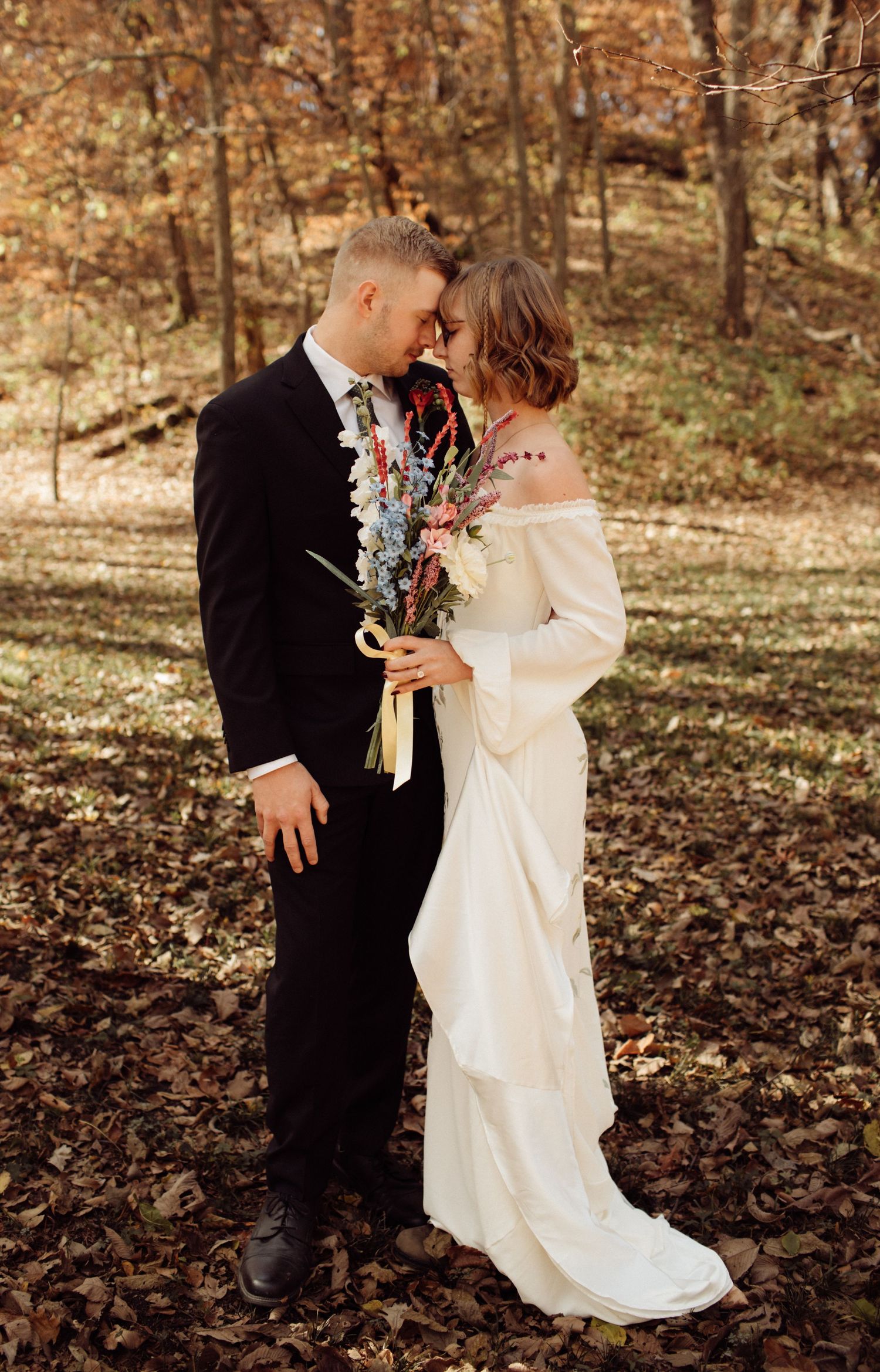 Bride and groom embracing in autumn forest, bride holding wildflower bouquet in embroidered gown.