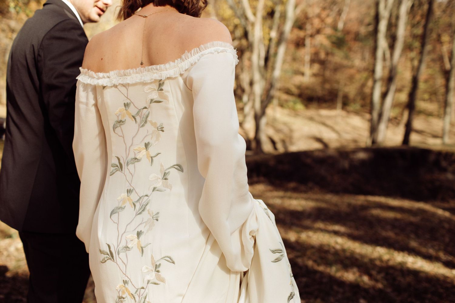 Bride in off-shoulder white gown with floral embroidery walking with groom in autumn forest.