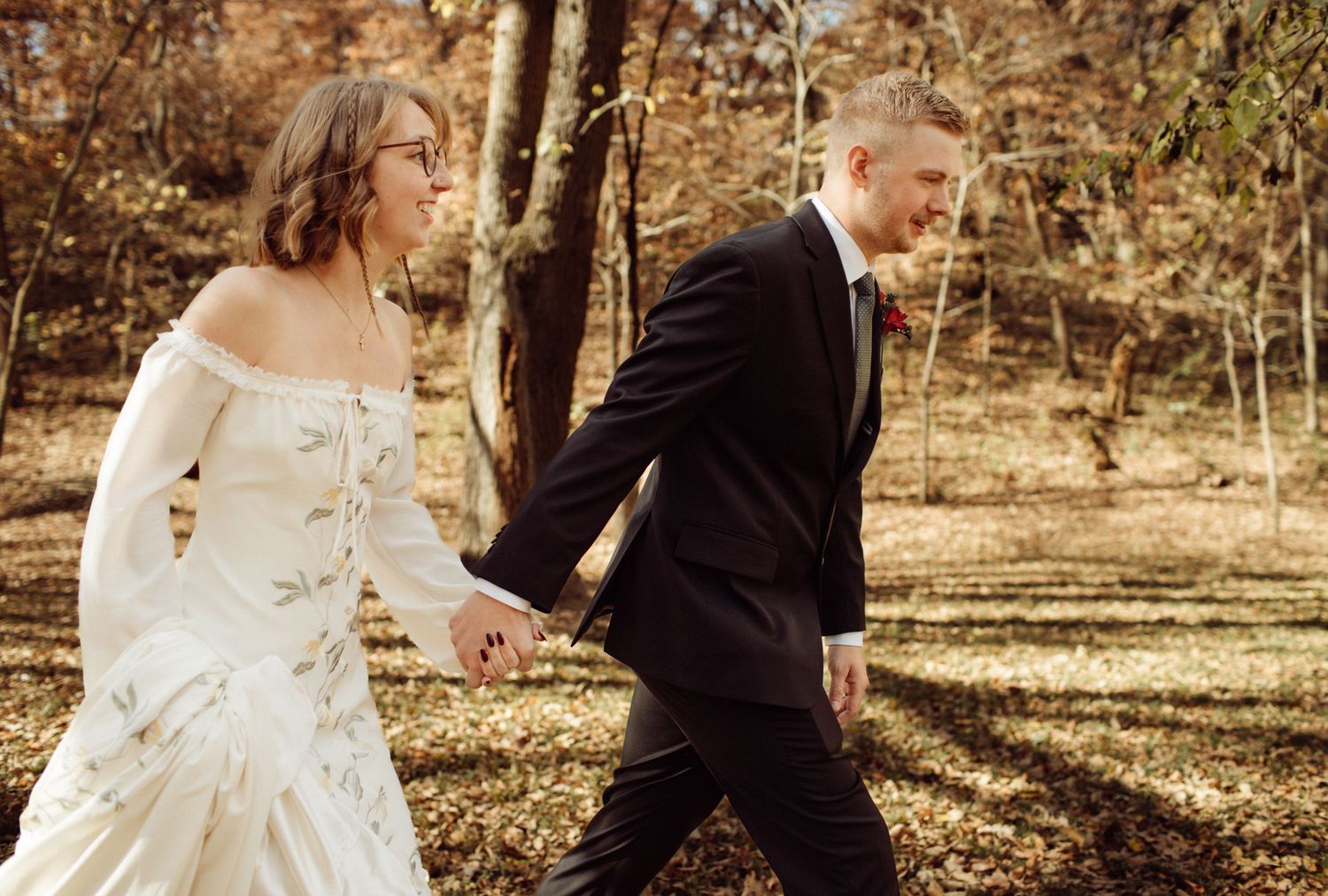 Smiling bride and groom holding hands walking through sunlit autumn forest on wedding day.