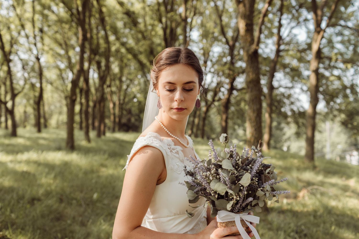 Elegant bride in white lace gown holding wildflower bouquet with eyes closed in sunlit forest setting.