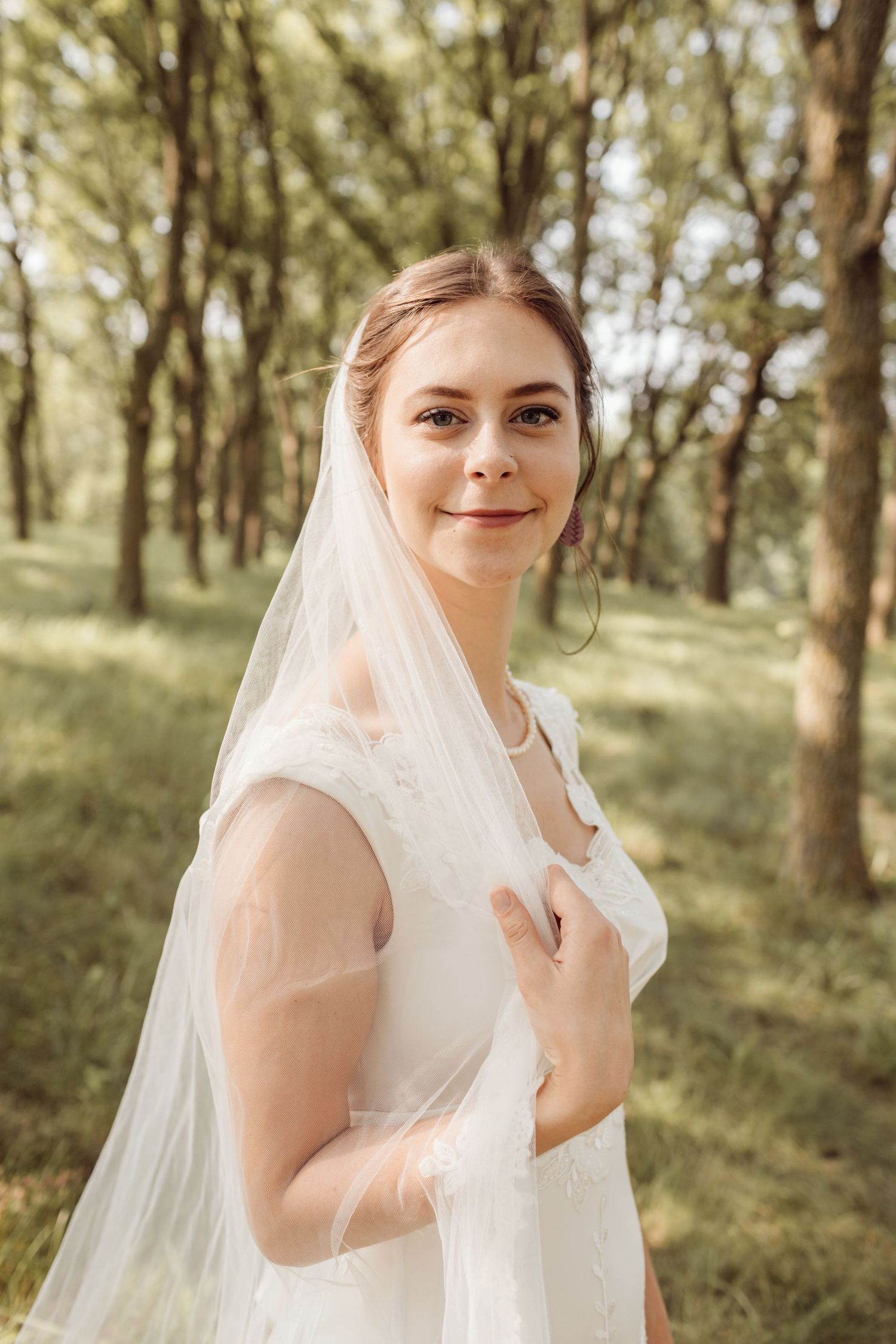 Beautiful bride in white wedding dress and veil smiling softly in sunlit tree-lined outdoor setting.