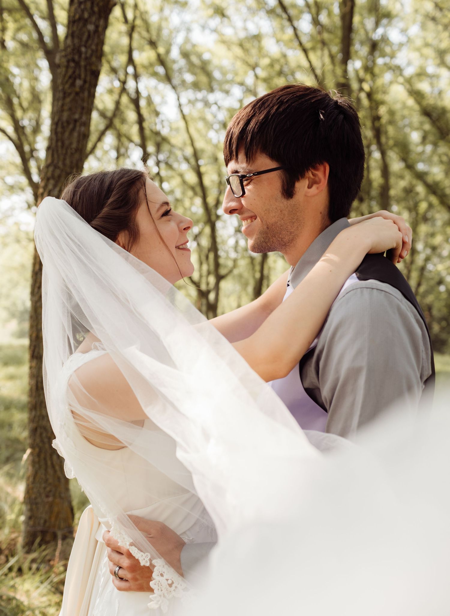 Groom lifts bride in white wedding dress and veil among green trees in romantic outdoor ceremony photo.