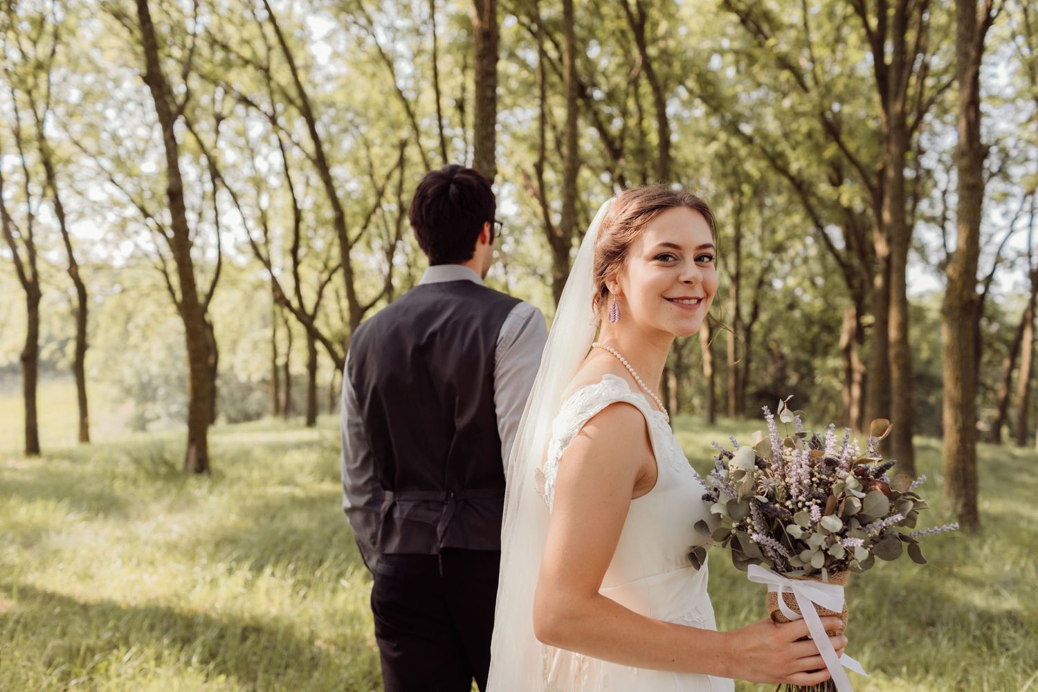 Bride in white gown holds bouquet and smiles over shoulder as groom stands in sunlit forest setting.