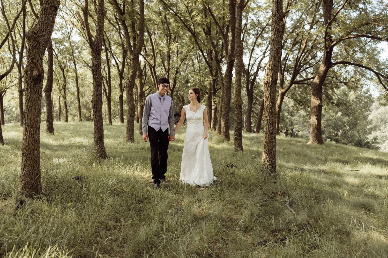 Bride and groom walk hand in hand through lush green woodland in elegant wedding attire.