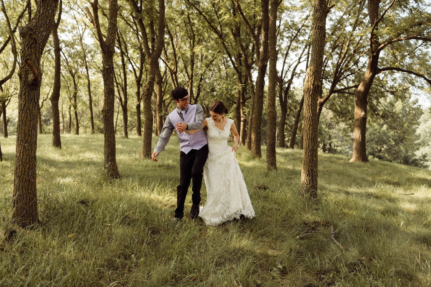 Joyful bride and groom run playfully through sunlit forest during outdoor wedding photo session.