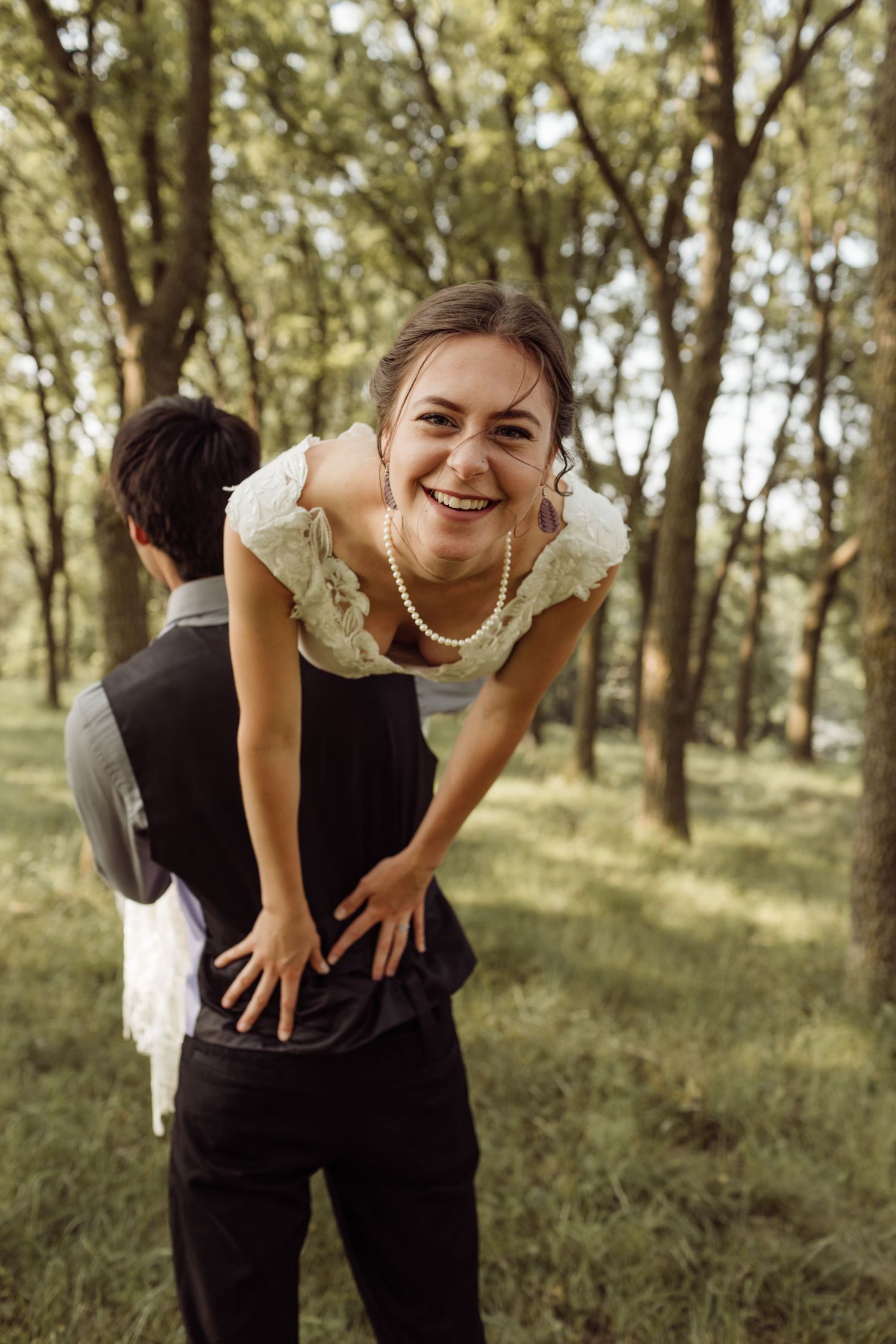 Laughing bride in lace top and pearl necklace leans forward during fun outdoor wedding piggyback moment.