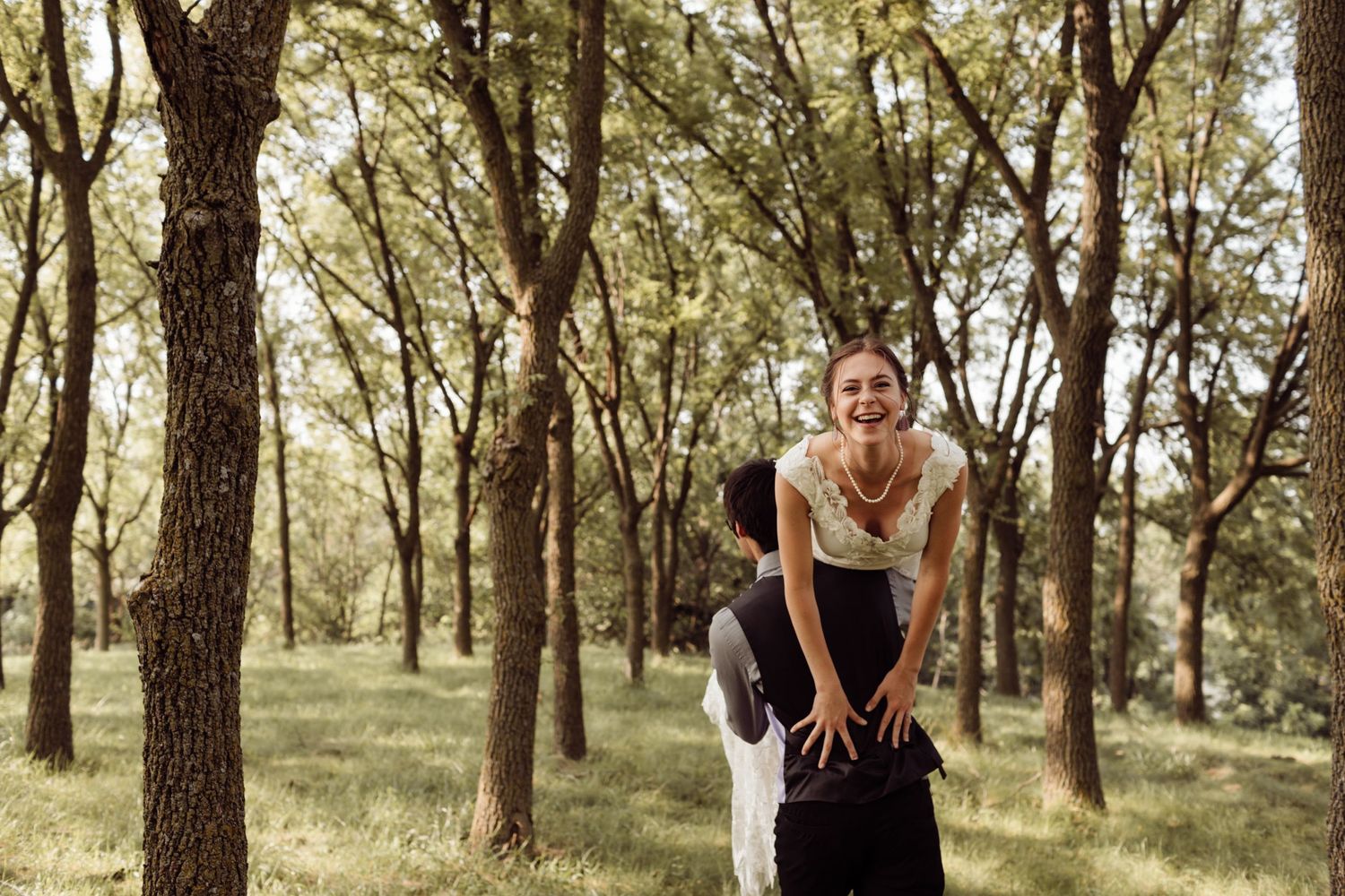 Groom lifts smiling bride in white lace dress during playful outdoor wedding photo in green forest.