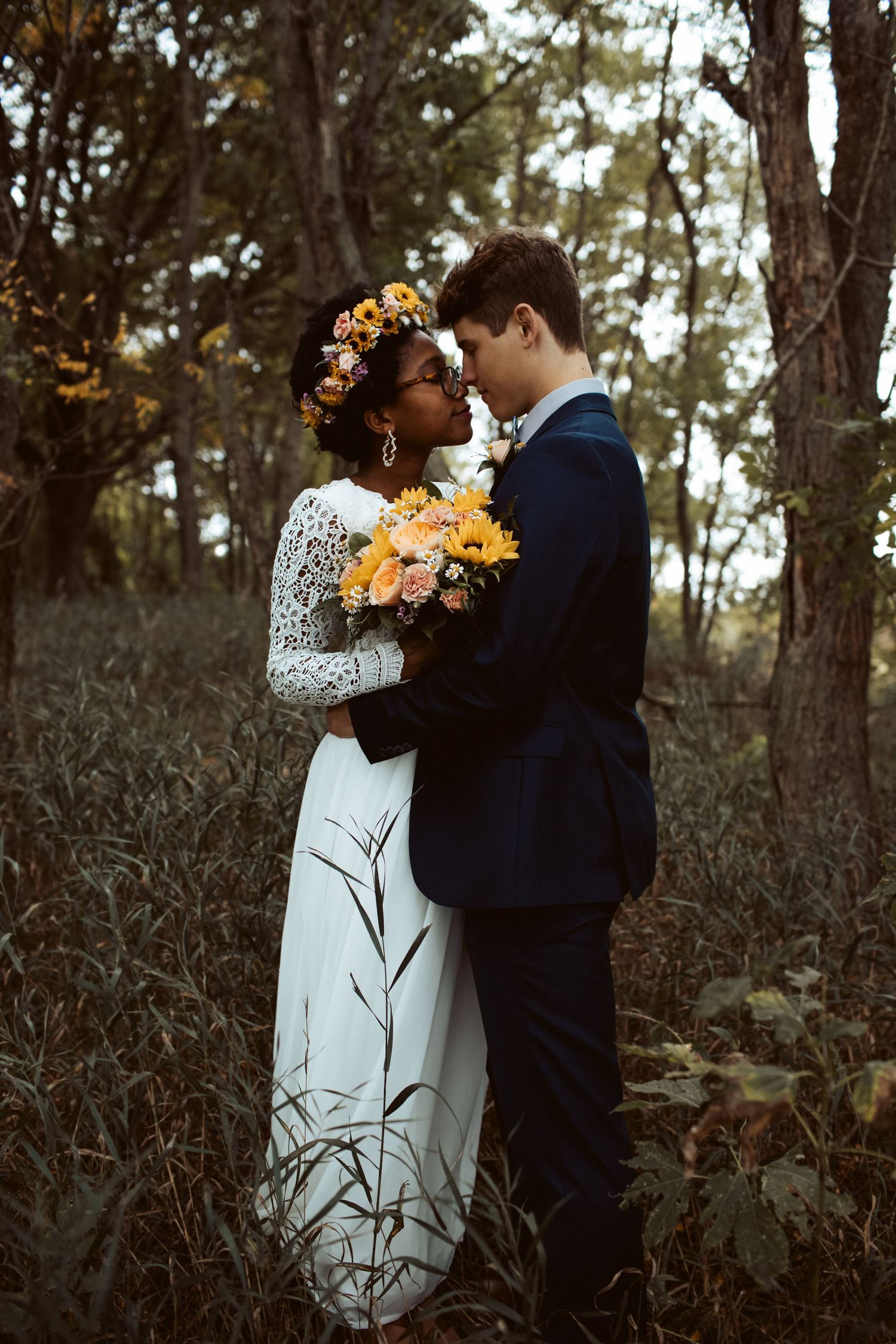 Couple in wedding attire embrace closely holding sunflower bouquet in moody forest setting.