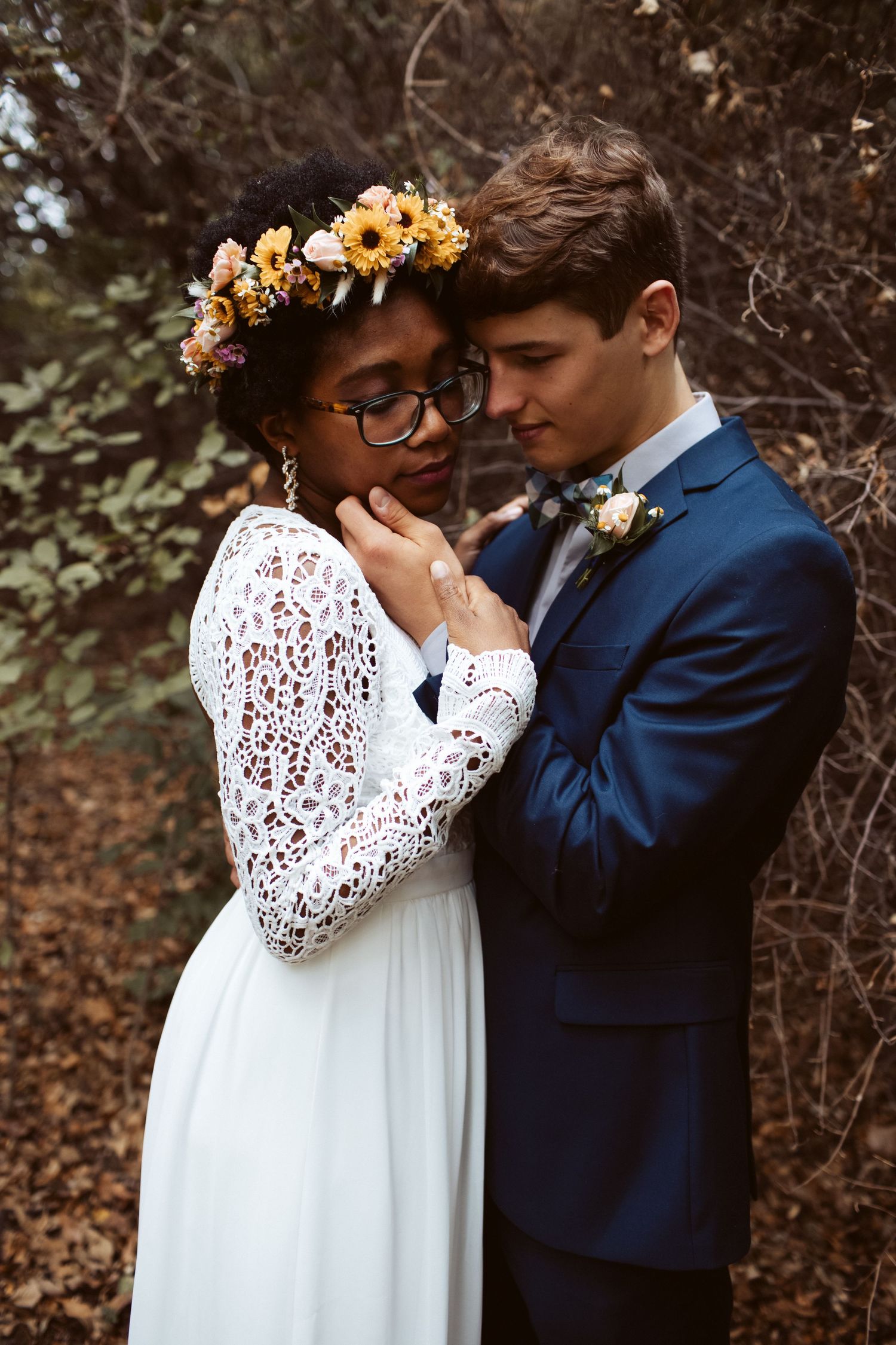 Bride in lace dress with flower crown leans into groom in navy suit during intimate forest wedding.