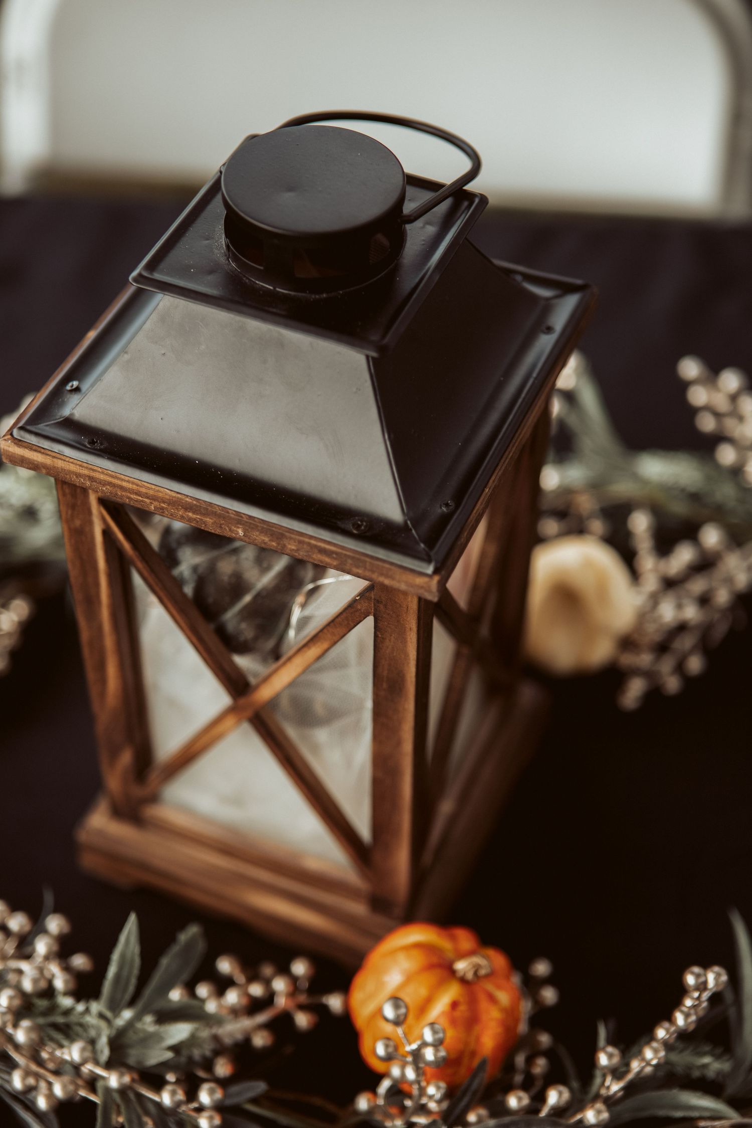 Rustic wooden lantern centerpiece with mini pumpkin, silver berries, and greenery on dark table.