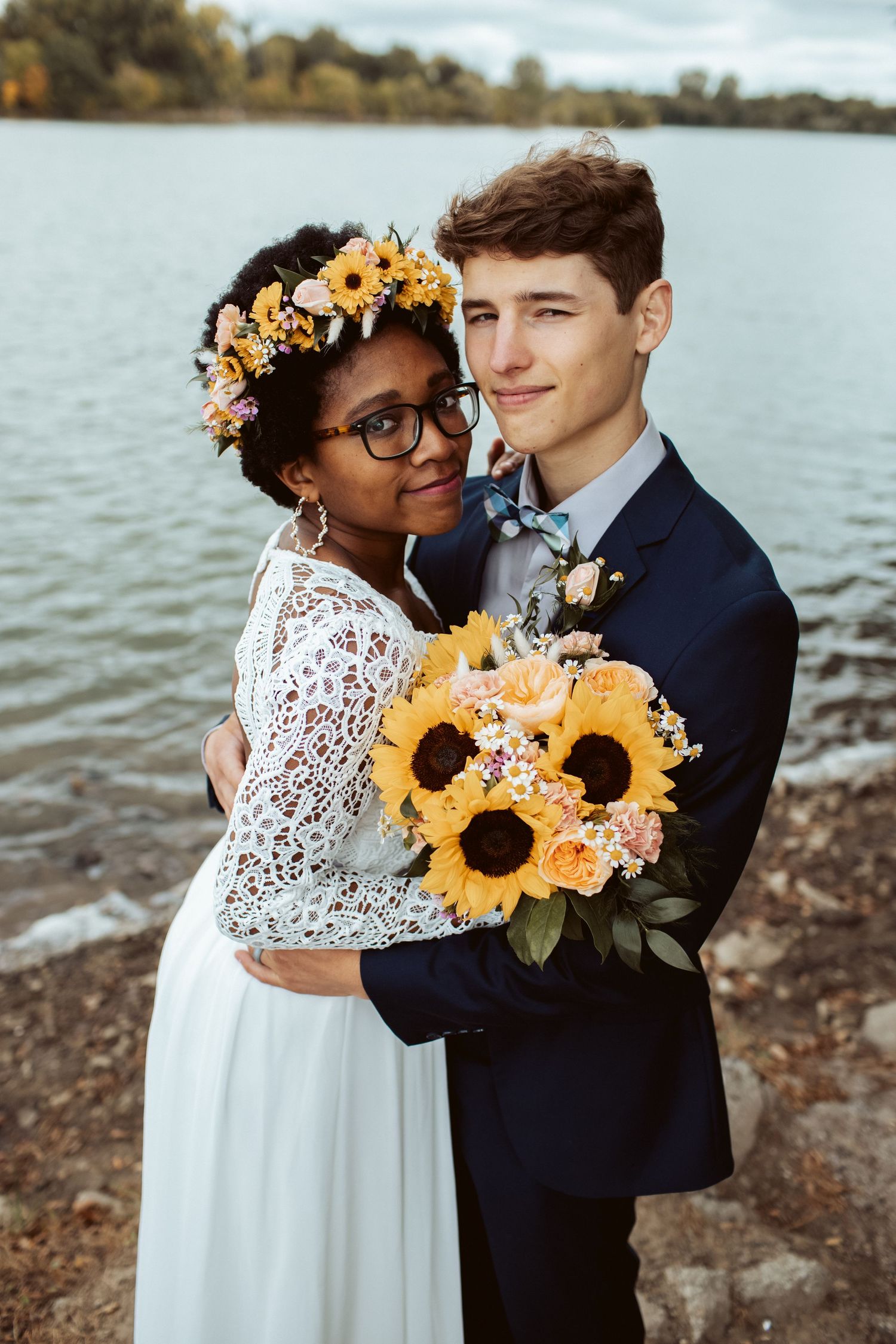Newlyweds embrace holding sunflower bouquet with flower crown bride near lakeside in fall.