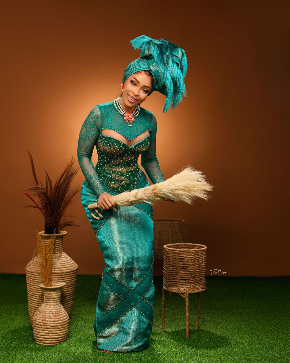 Elegant figure in turquoise asa oke attire with feathered headpiece and fan poses against brown backdrop on artificial grass.